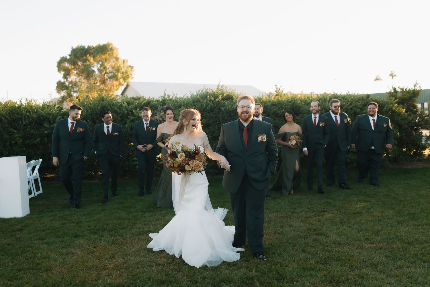 Wedding party walking across green lawn with bride and groom leading groomsmen and bridesmaids.