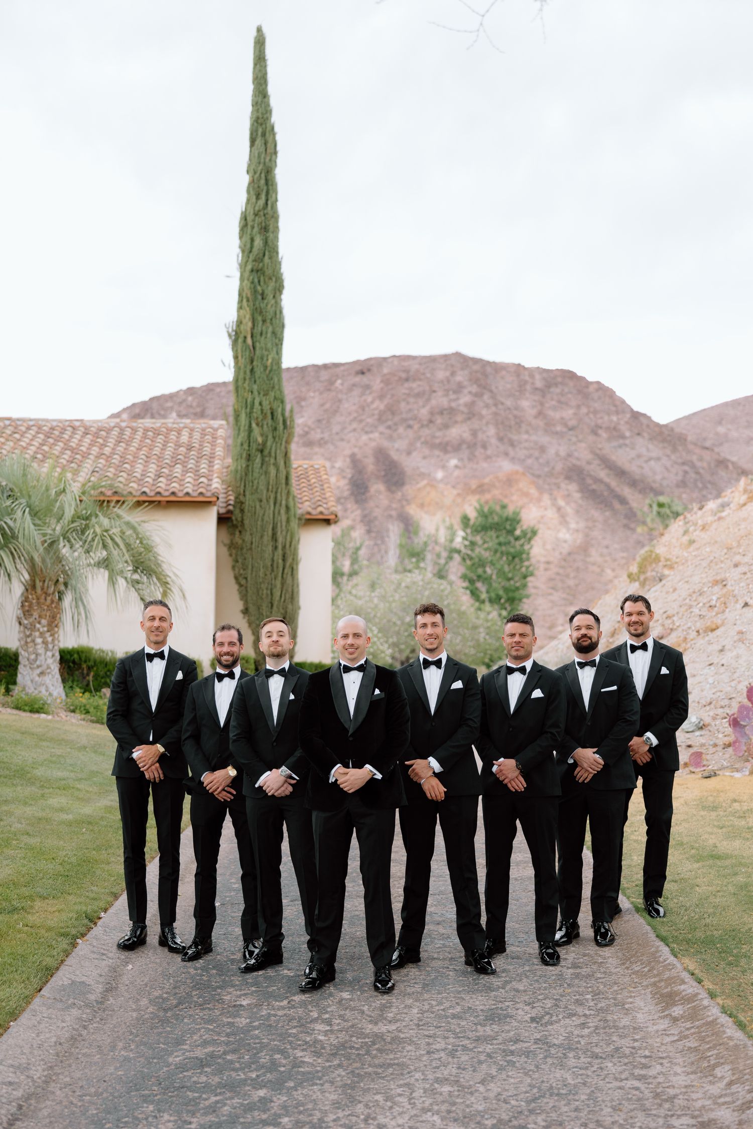Groom and groomsmen in black tuxedos pose on pathway with cypress tree and mountain backdrop.