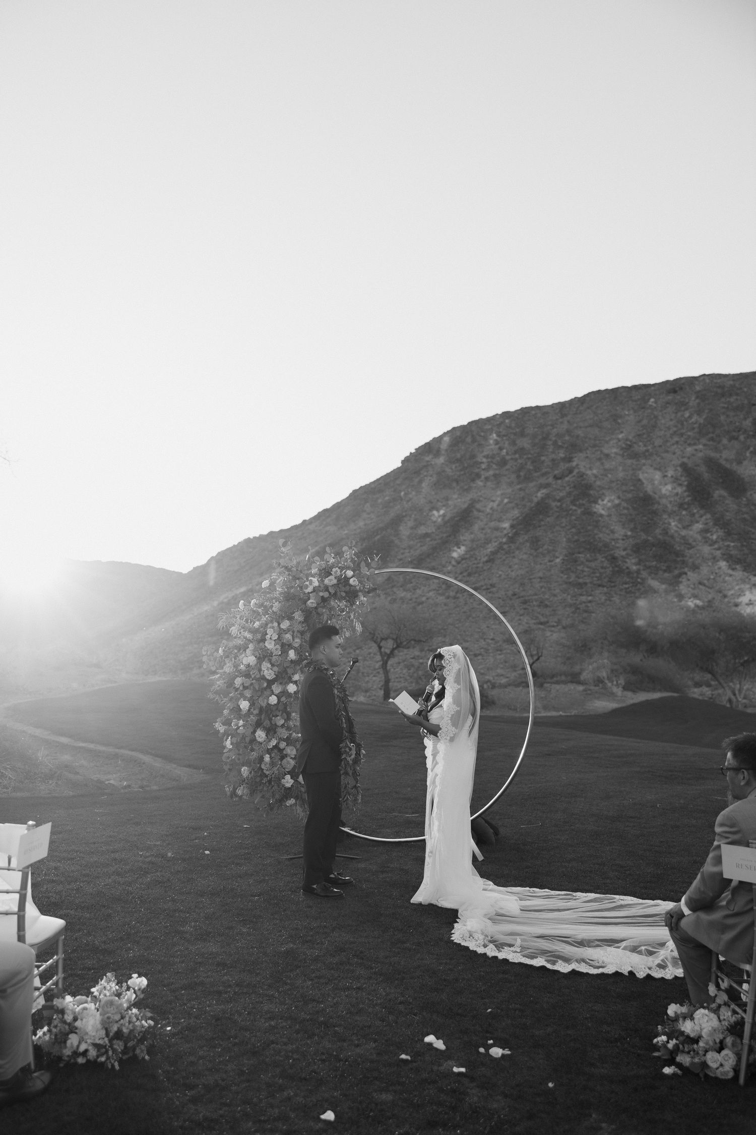 Black and white photo of bride and groom exchanging vows under a circular floral arch with mountain backdrop.