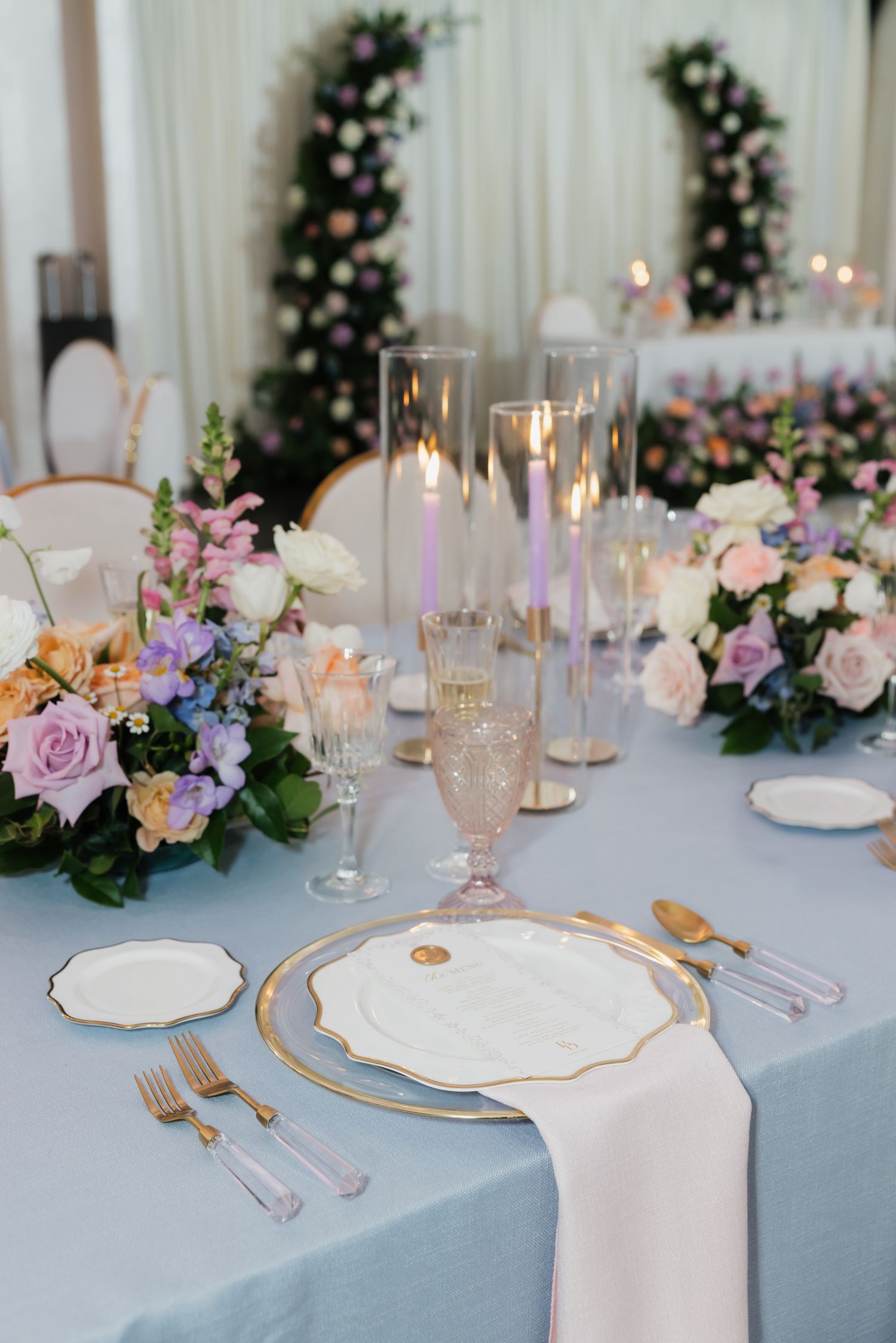 Elegant wedding table setting with blue linen, gold cutlery, white plates, purple candles, and pastel floral arrangements.