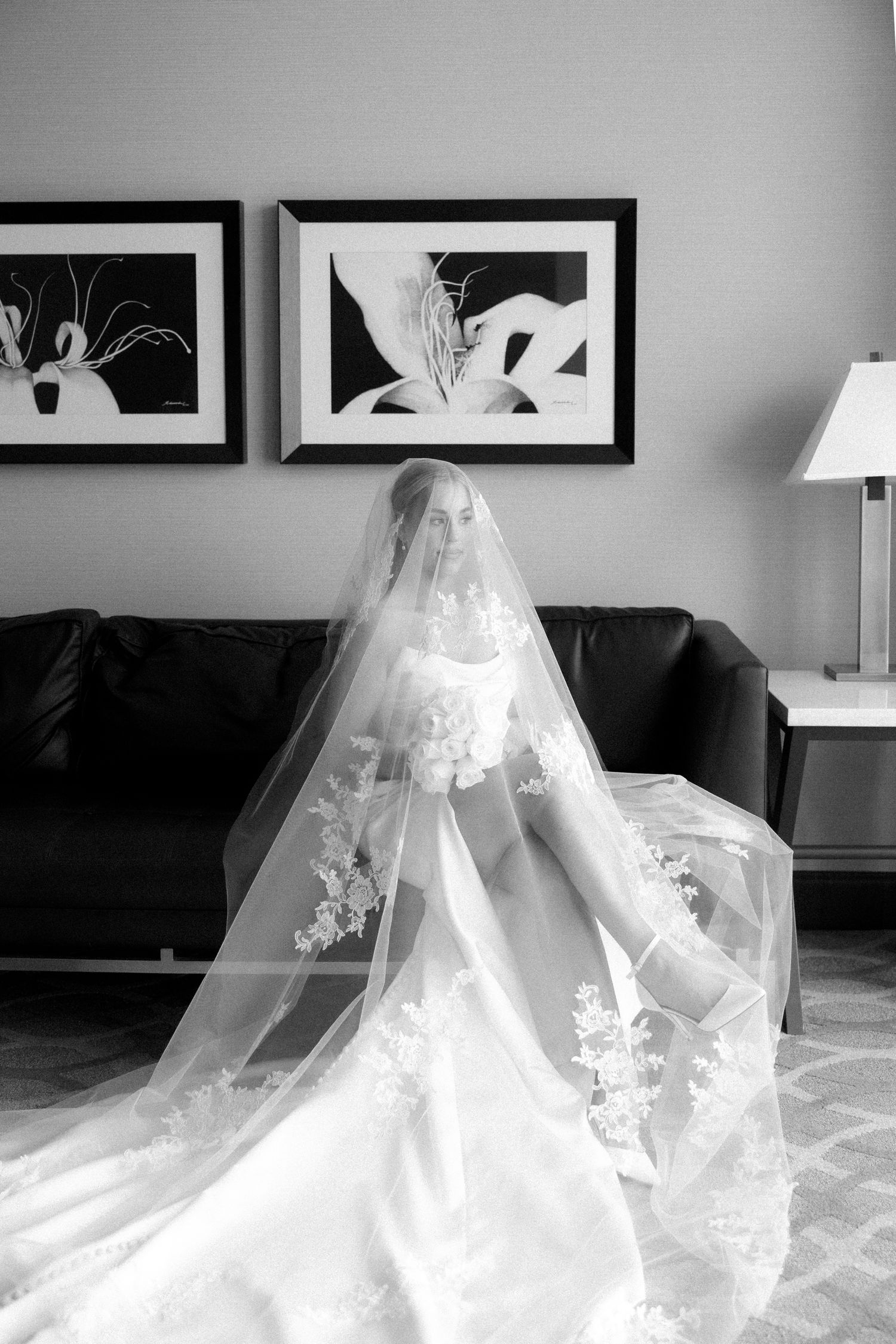 Bride in elegant lace wedding gown and long cathedral veil seated in black and white portrait photo.