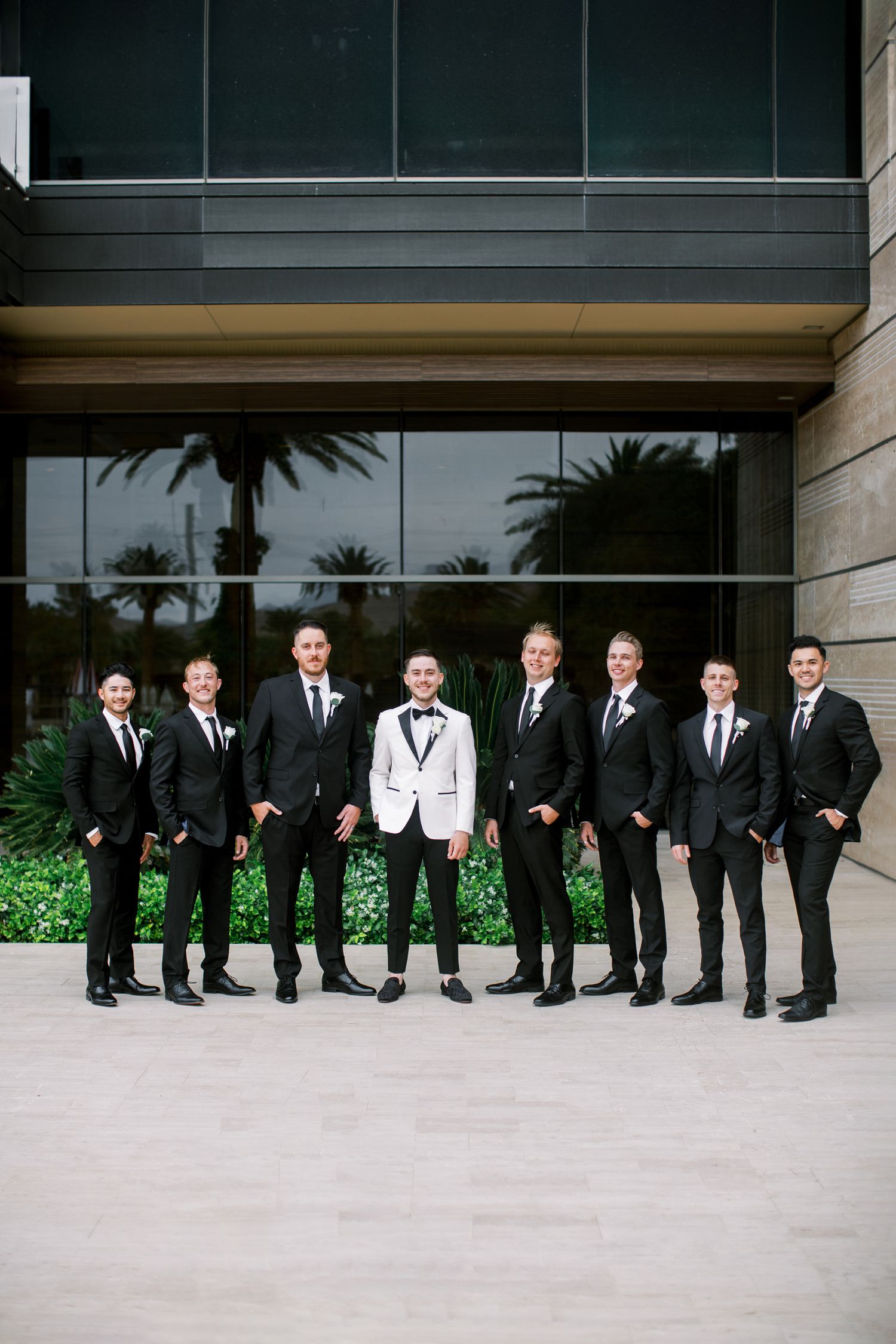 Groom in white tuxedo jacket poses with groomsmen in black suits outside modern glass building.