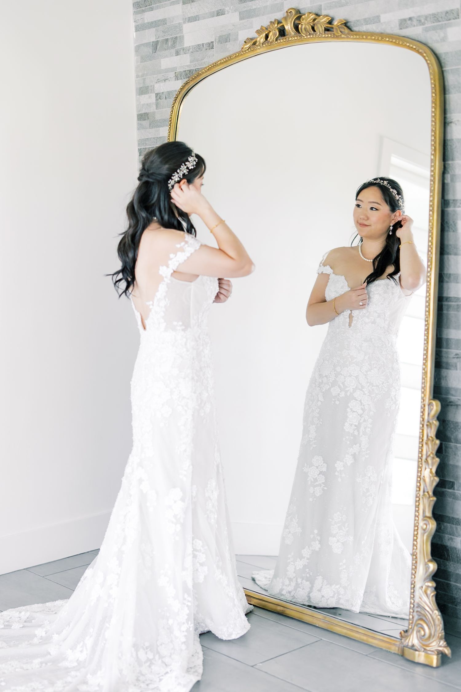 A bride in a flowing white lace wedding dress admires her reflection in an ornate gold-framed mirror.