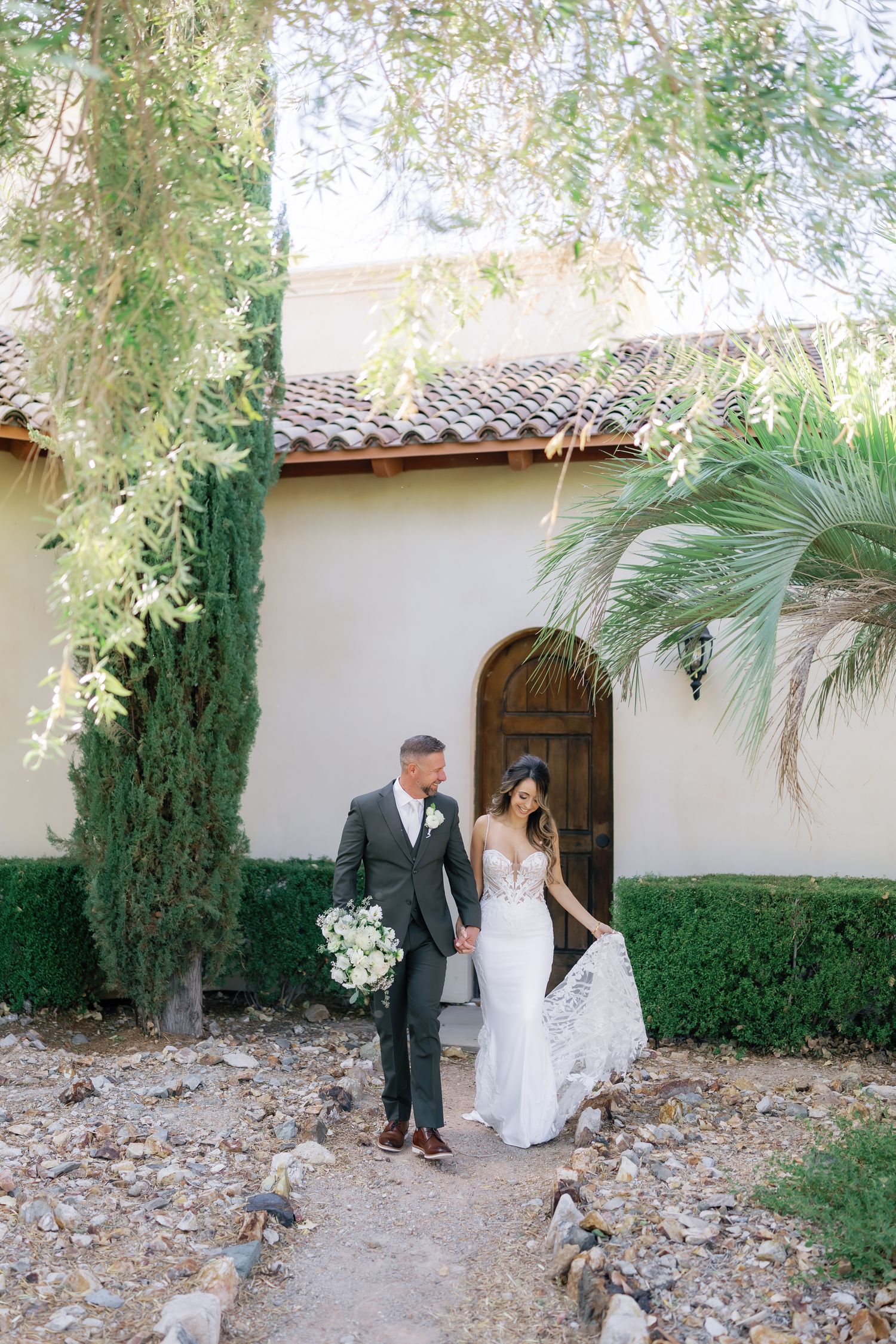 A couple stands in a Mediterranean-style doorway with white stucco walls, cypress trees and palm fronds framing the scene.