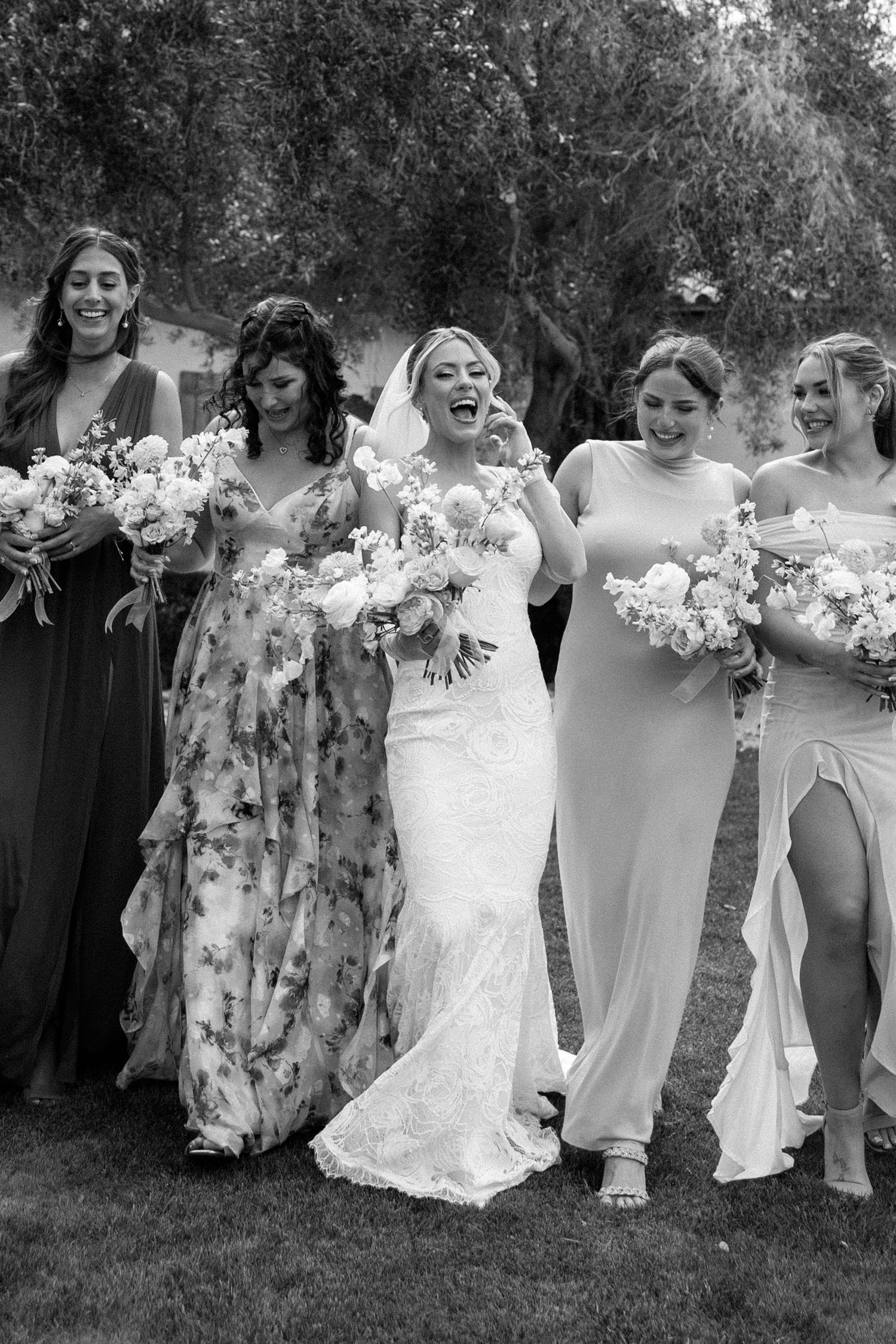Black and white photo of a laughing bride in lace gown walking with five bridesmaids holding bouquets outdoors.