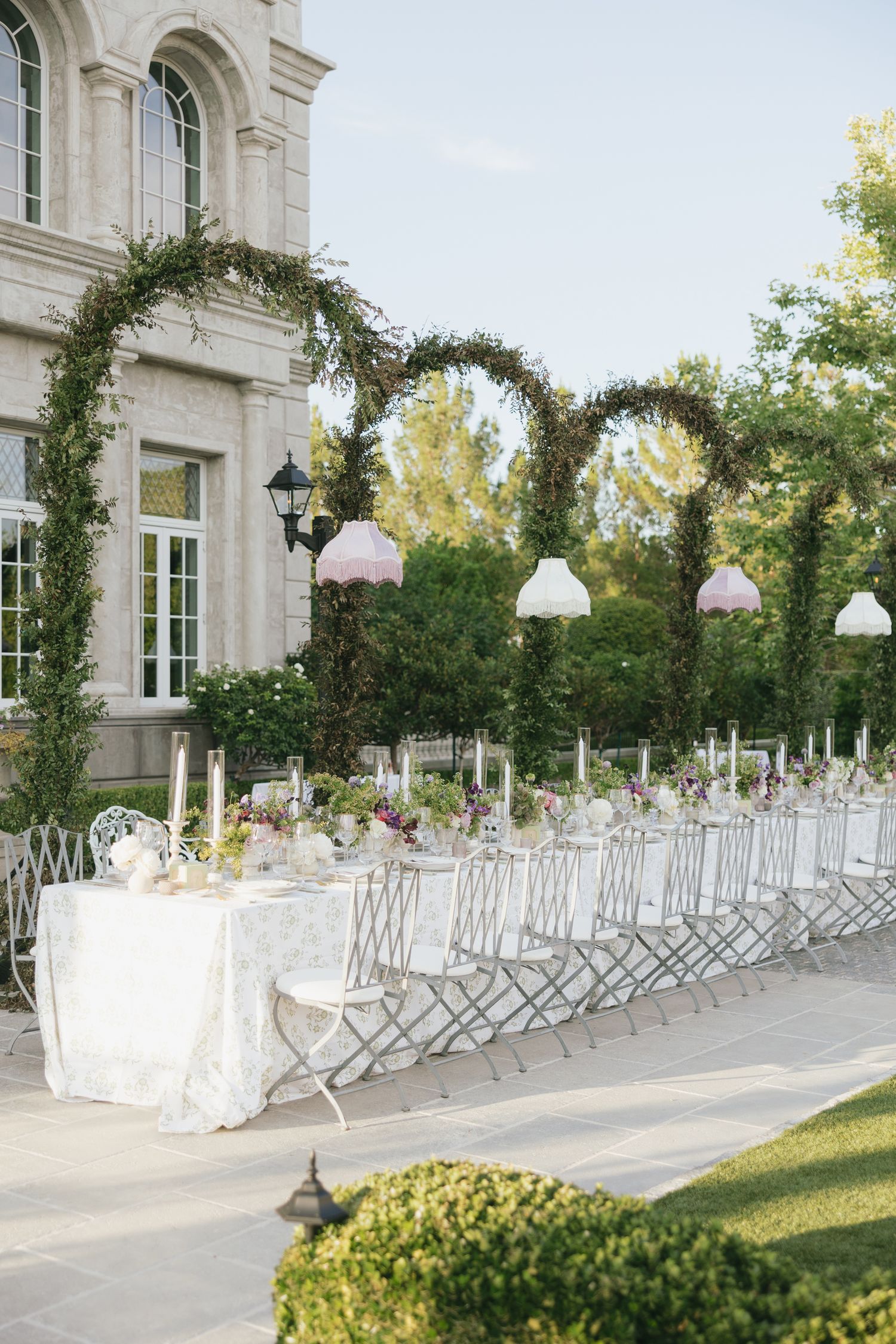 Elegant outdoor wedding reception with long table, floral arches, pink pendant lights, and white chairs beside a grand stone building.