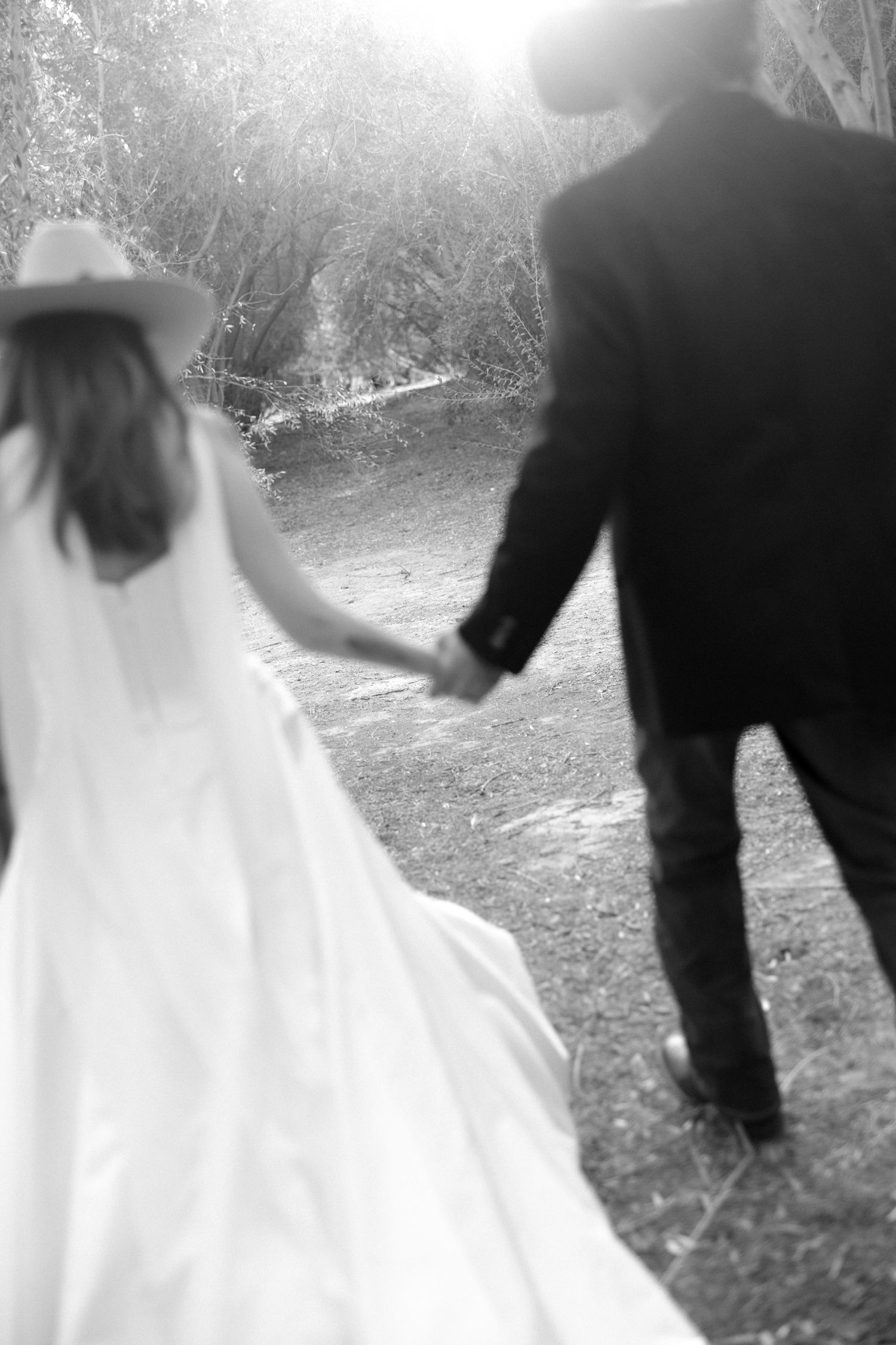 Black and white photo of bride in white gown and cowboy hat holding hands with groom in suit walking through nature.