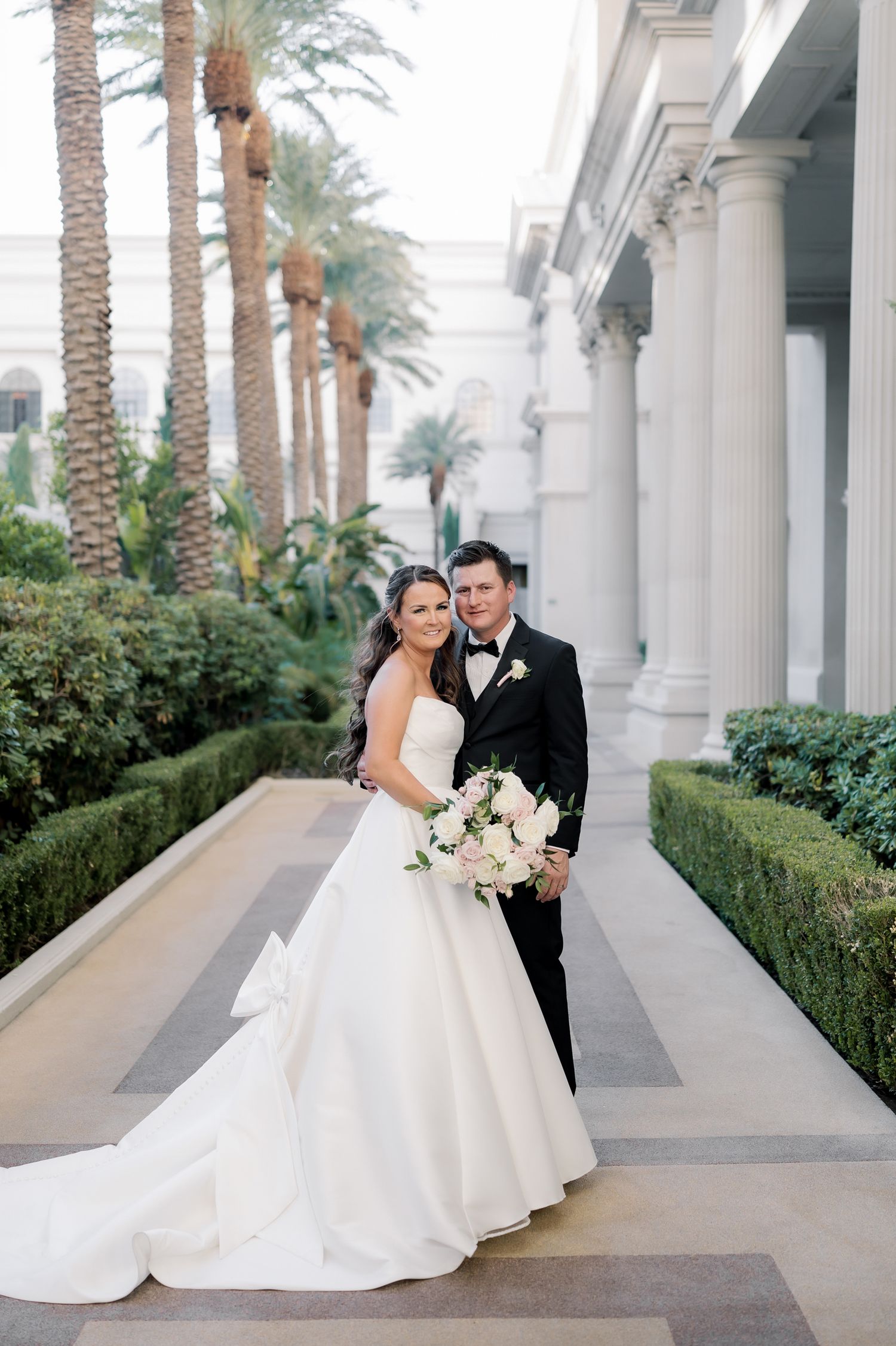 A romantic wedding couple poses together in an elegant colonnade walkway lined with palm trees and greenery.