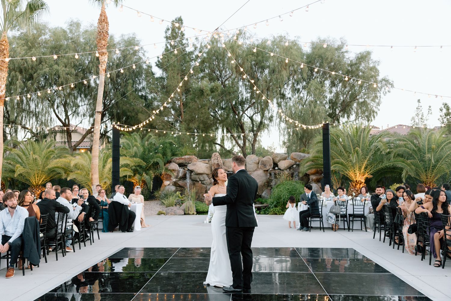 Couple dancing on an elevated white platform during an outdoor evening reception with guests seated around them.