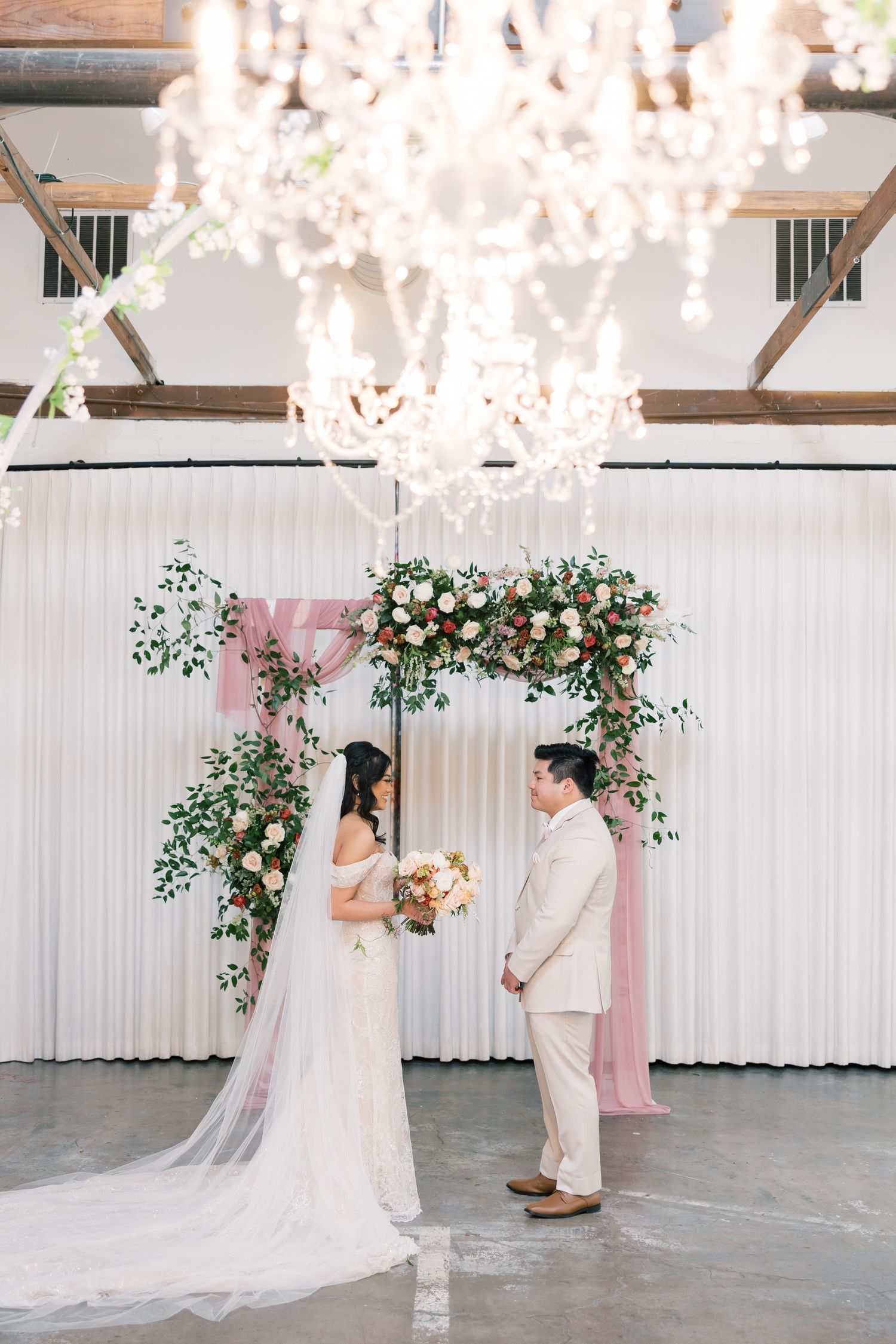 Wedding ceremony setup with floral arch, white draping, and crystal chandeliers in an industrial venue.