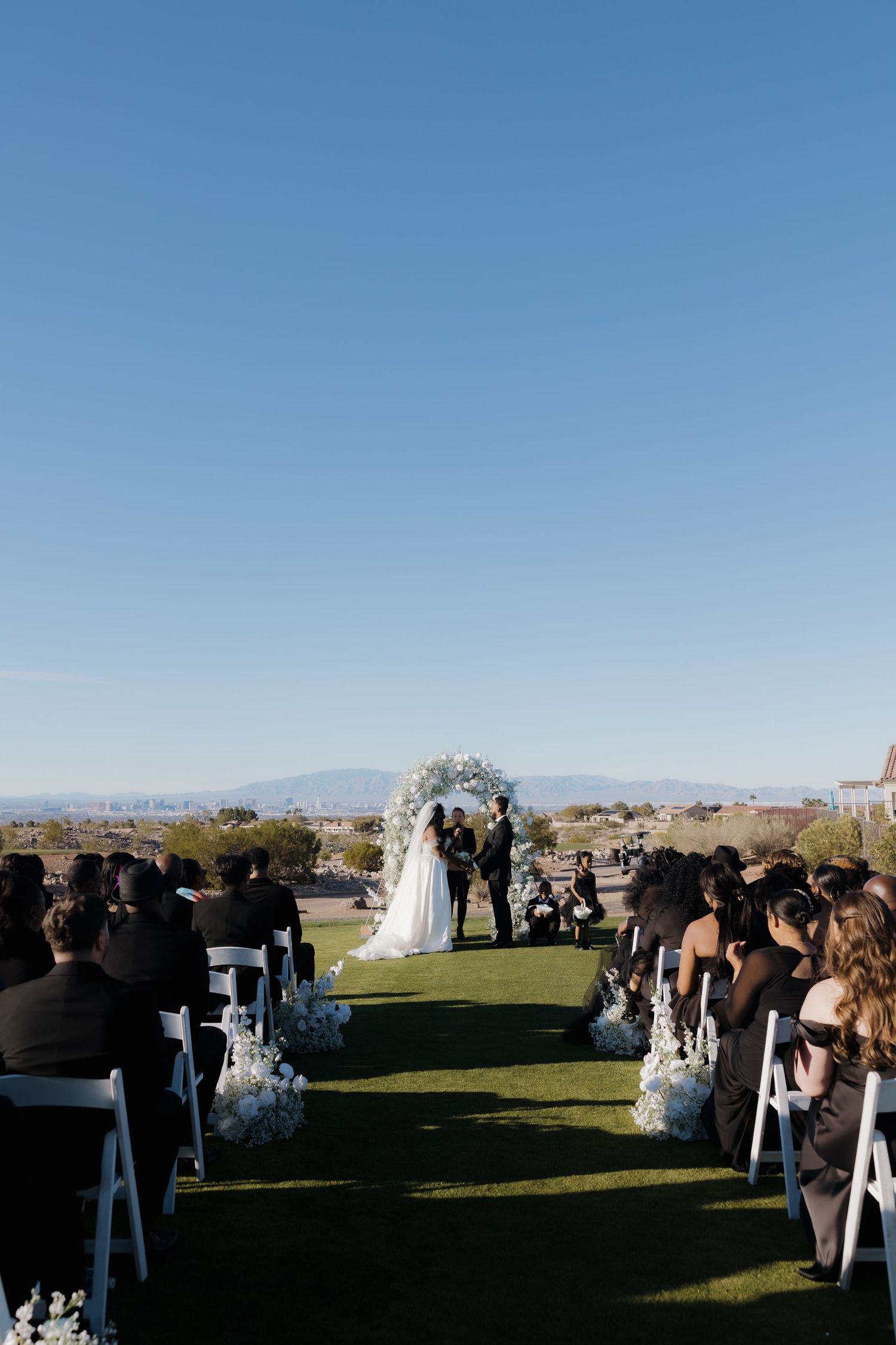 Outdoor wedding ceremony on green lawn with floral arch, guests in black attire seated in white chairs, mountain backdrop.