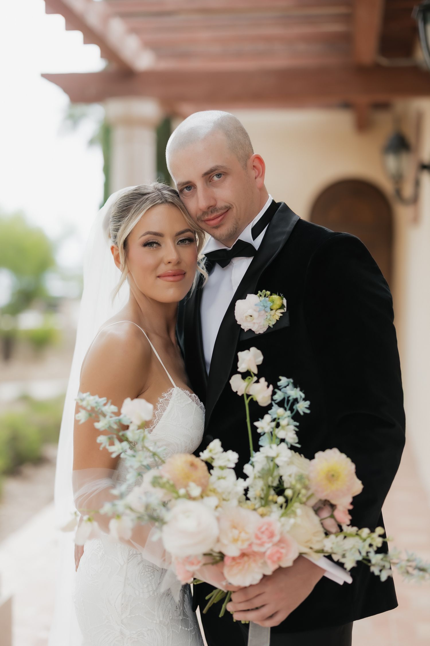 Bride holding blush floral bouquet poses with tuxedo-clad groom outside elegant venue with arched architecture.