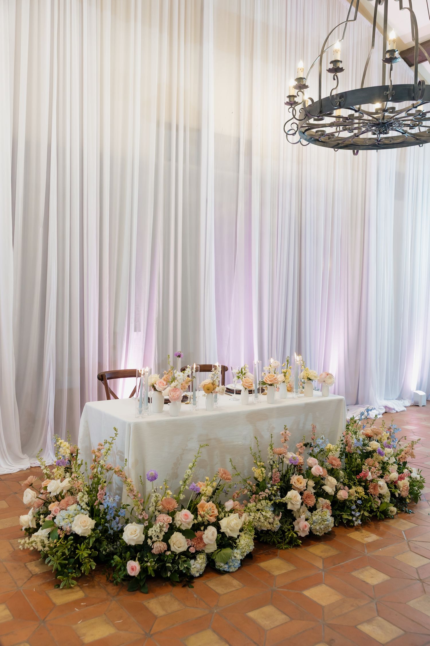 Elegant wedding sweetheart table adorned with lush pastel floral arrangements against sheer white draping.
