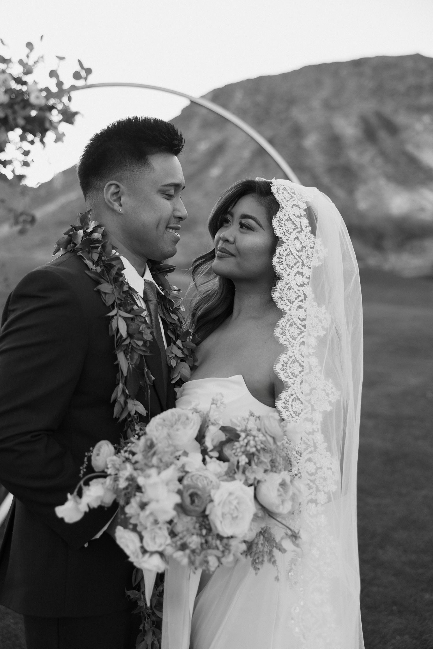 Black and white portrait of bride and groom at circular arch, bride in lace veil holding bouquet.