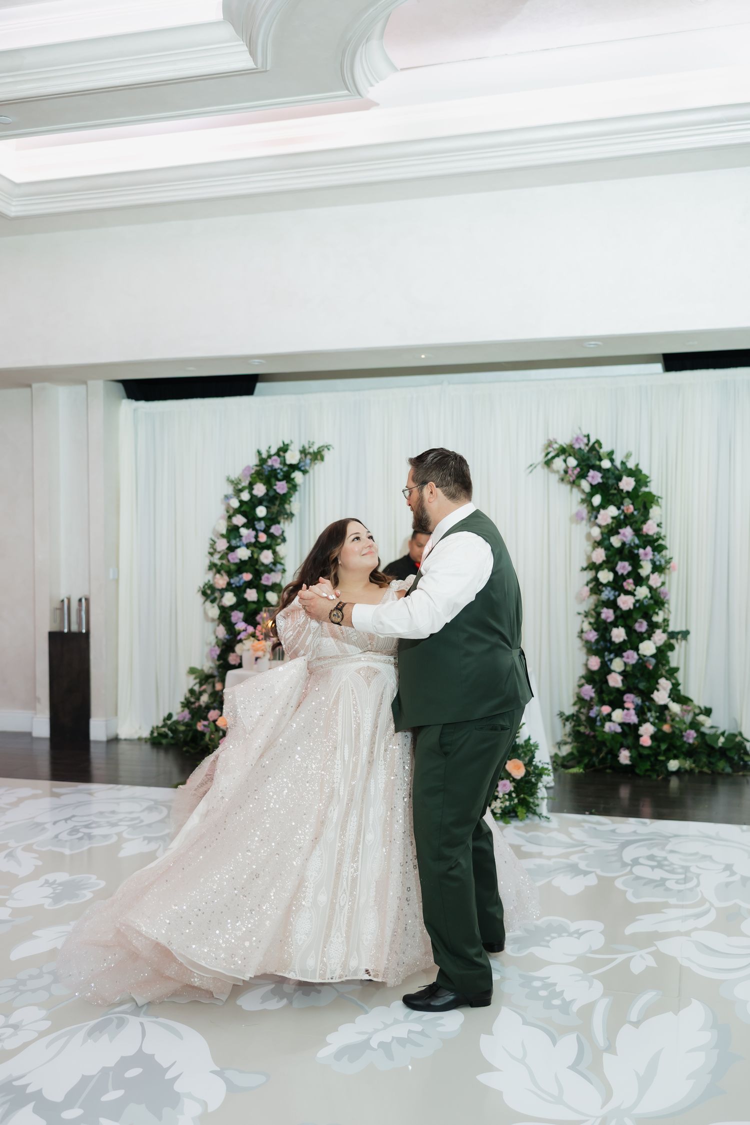Bride and groom share their first dance in an elegant ballroom with floral arch backdrop and decorative dance floor.