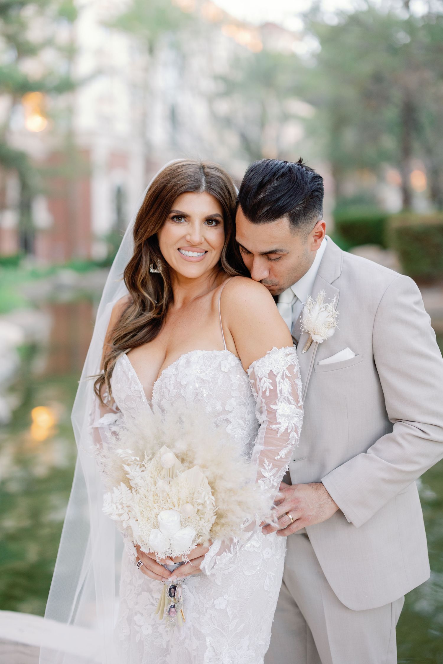 Bride in lace off-shoulder gown holds white bouquet as groom in tan suit kisses her cheek outdoors.