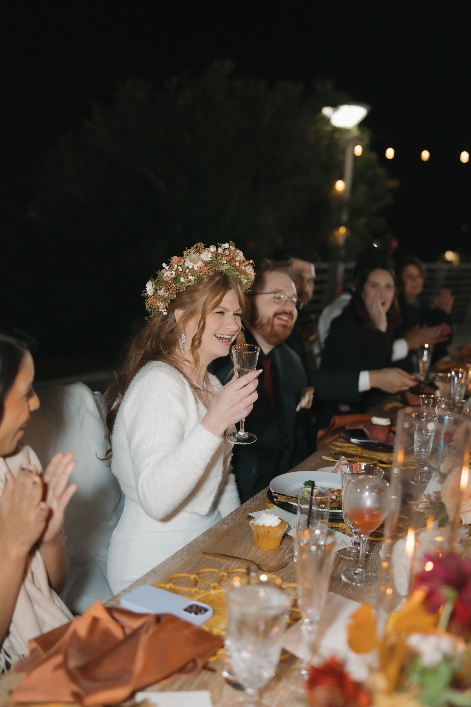 Bride wearing floral crown laughs while holding champagne glass at candlelit outdoor evening reception dinner.