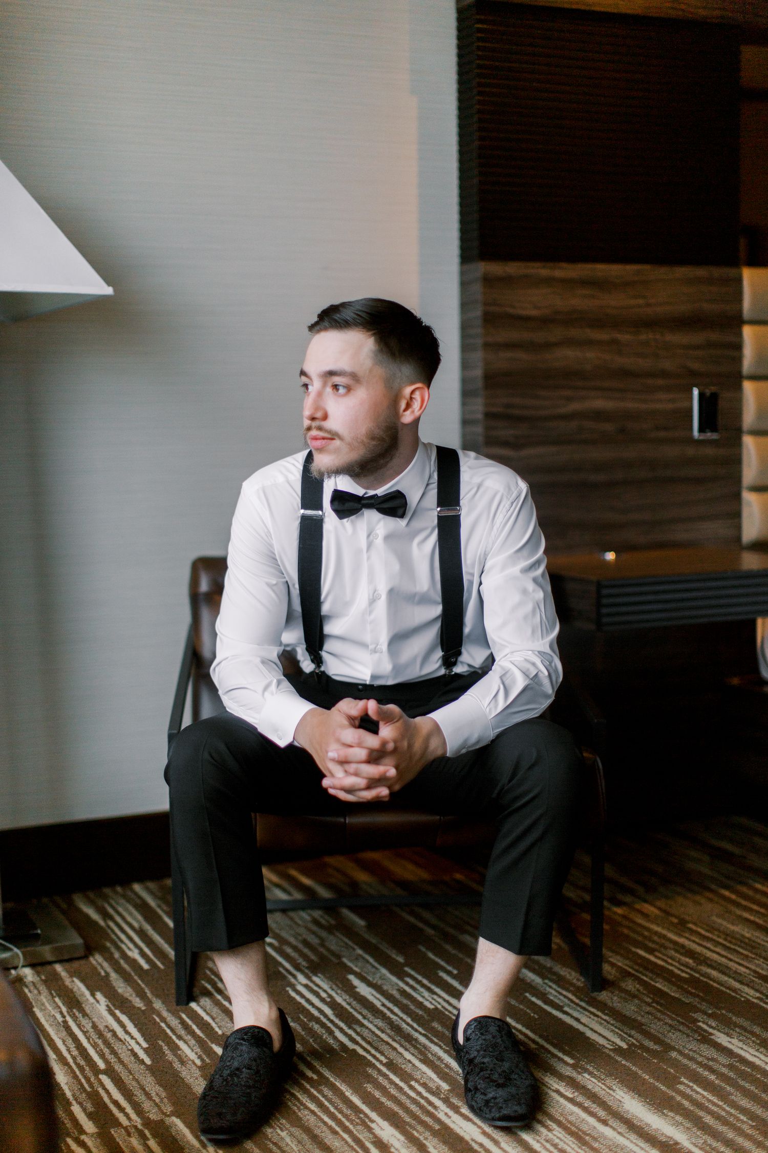 A dapper groom sits in a chair wearing a white shirt, black bow tie, suspenders, and dress shoes, gazing thoughtfully aside.