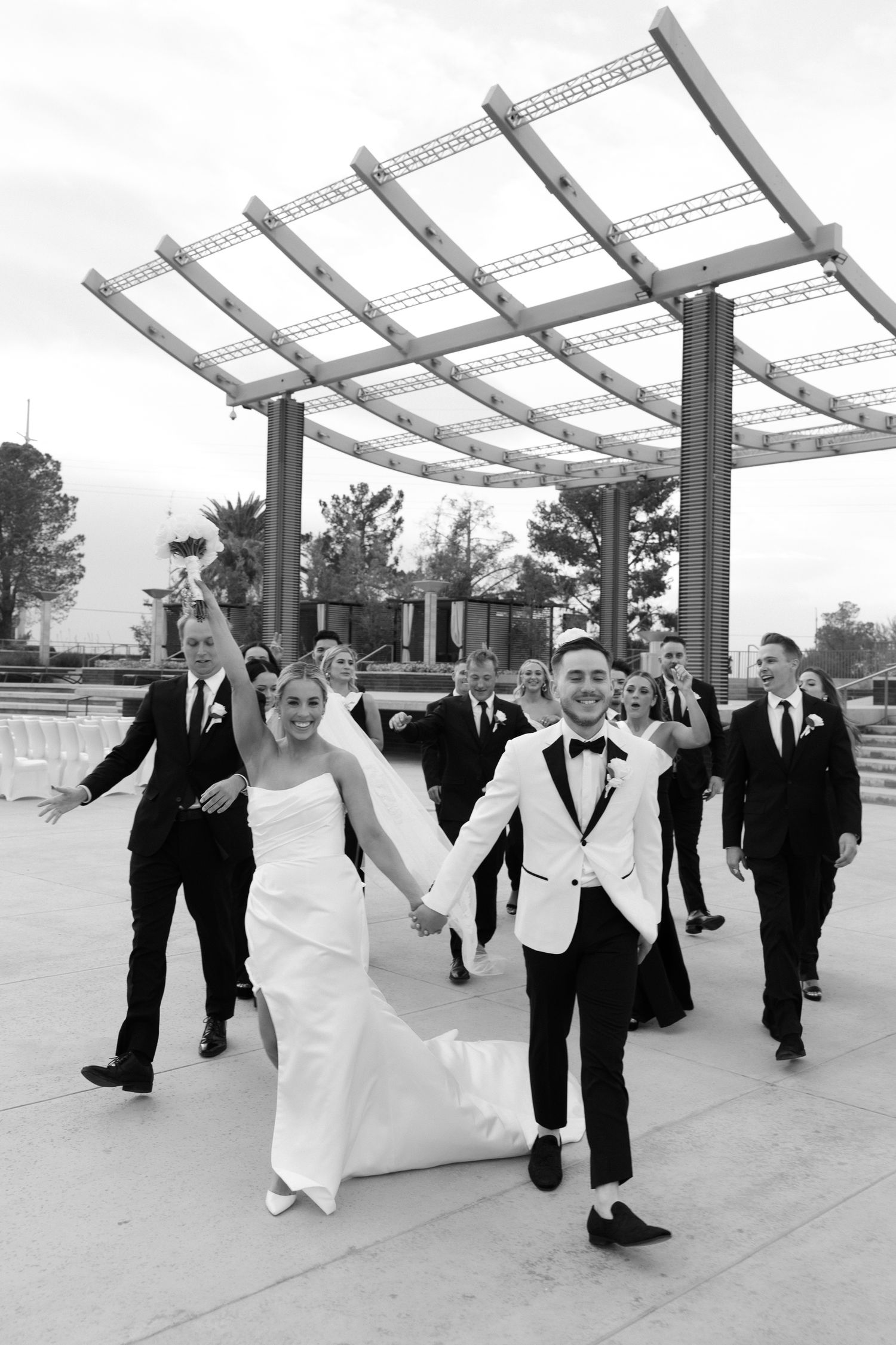Black and white photo of a joyful wedding party walking under a modern pergola after the ceremony.
