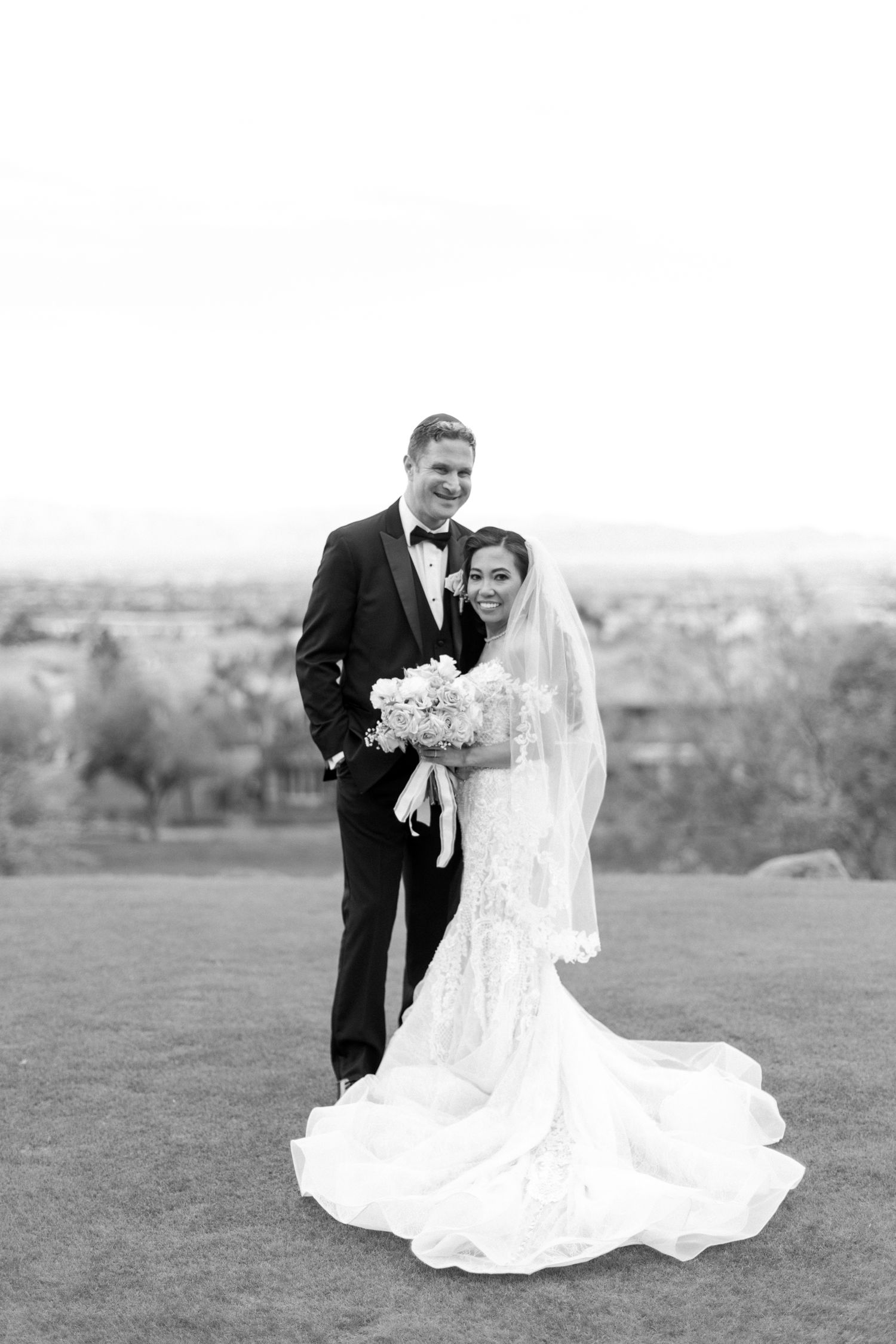 Black and white wedding portrait of smiling bride in lace gown with veil and groom in tuxedo standing on green lawn.