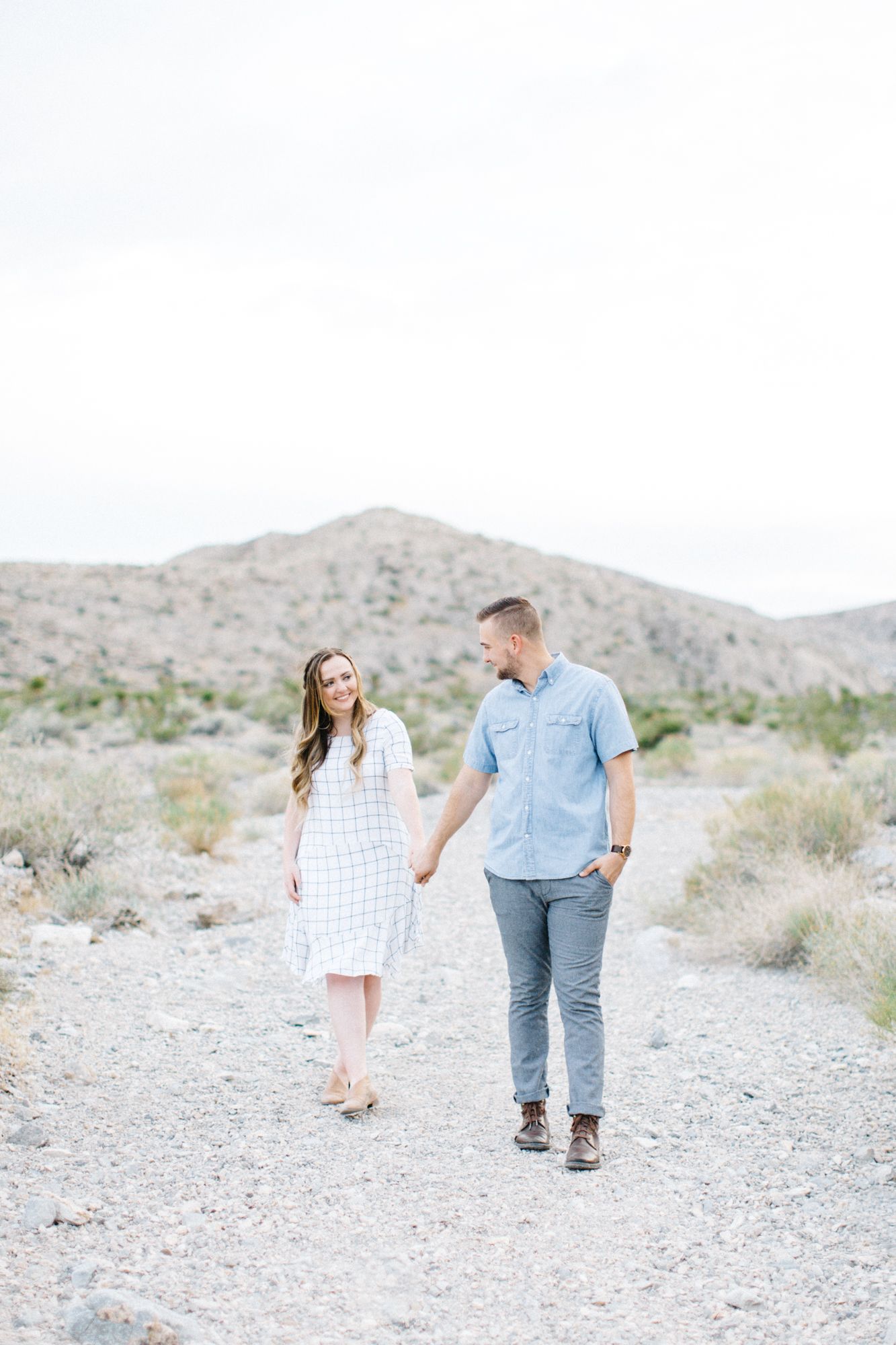 Couple holding hands walking on a desert trail with mountains in the background during an outdoor engagement session.