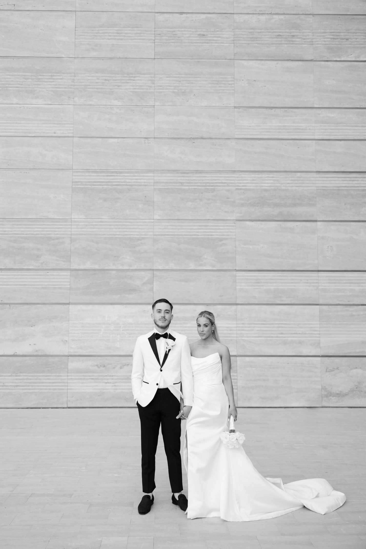 Black and white photo of bride and groom holding hands in front of a stone wall on their wedding day.