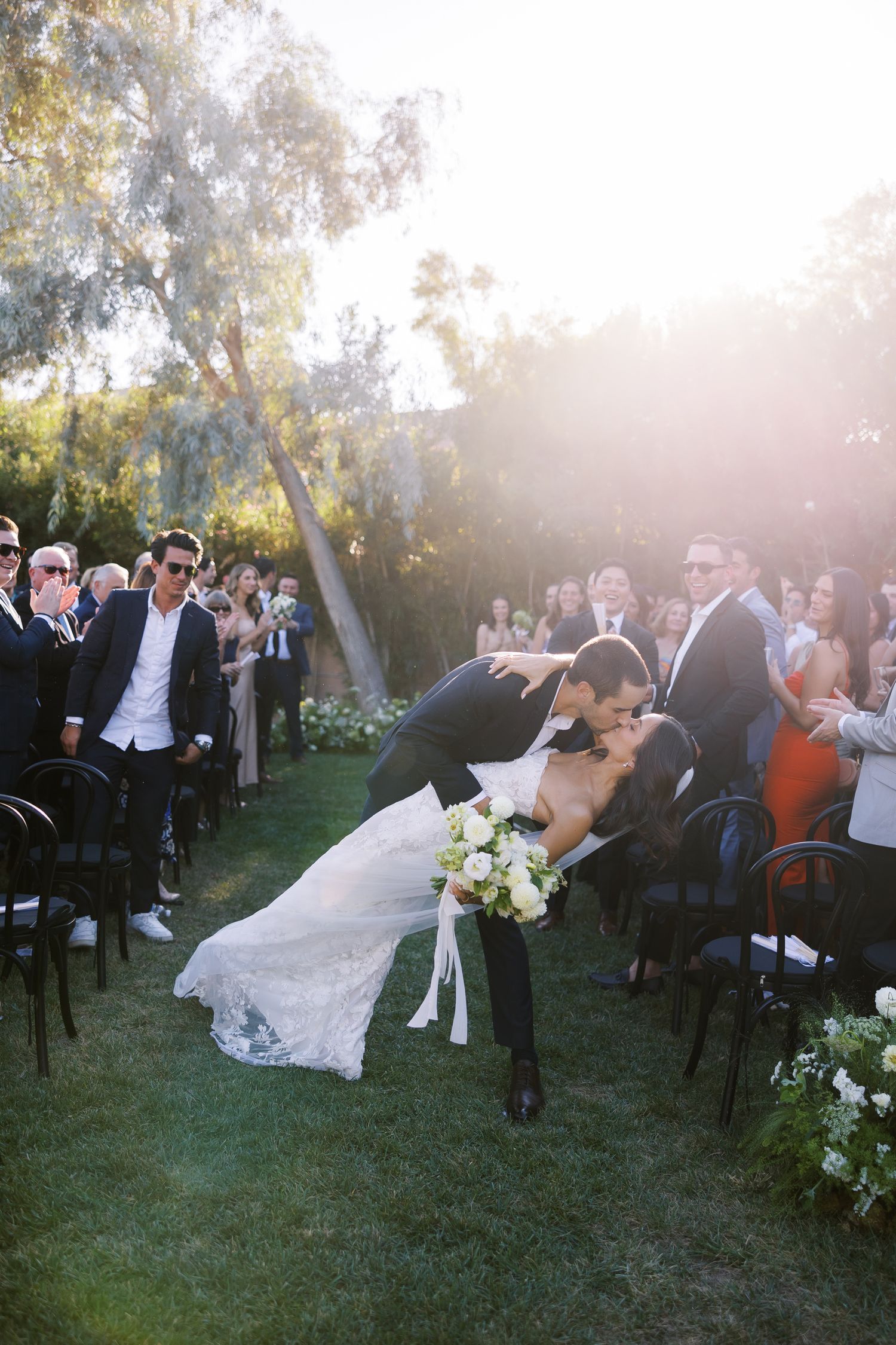 Groom dips bride in romantic outdoor wedding ceremony kiss, guests applauding, golden sunset light, lush garden setting.