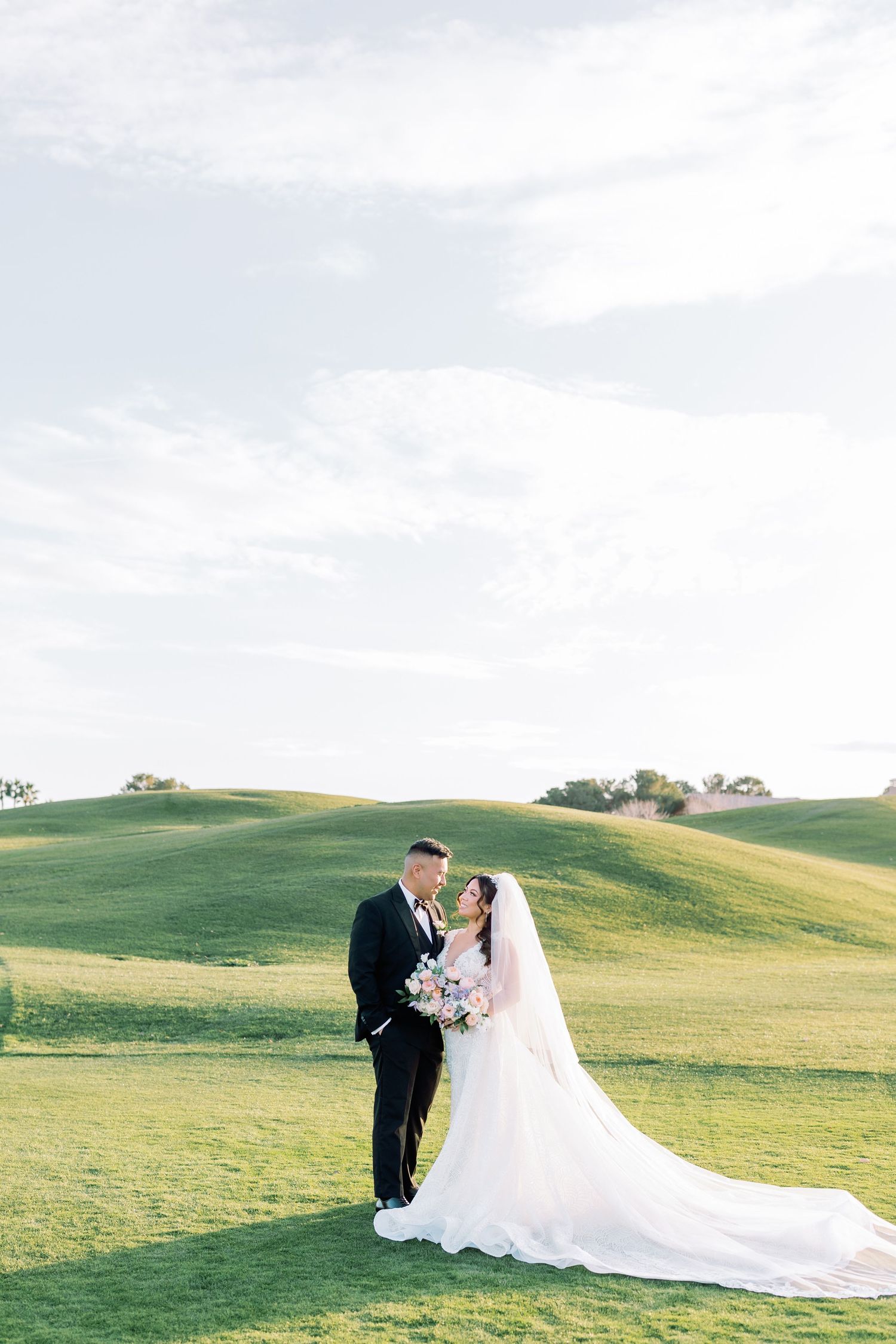 Bride and groom share a romantic moment on lush green golf course hills under a soft cloudy sky.