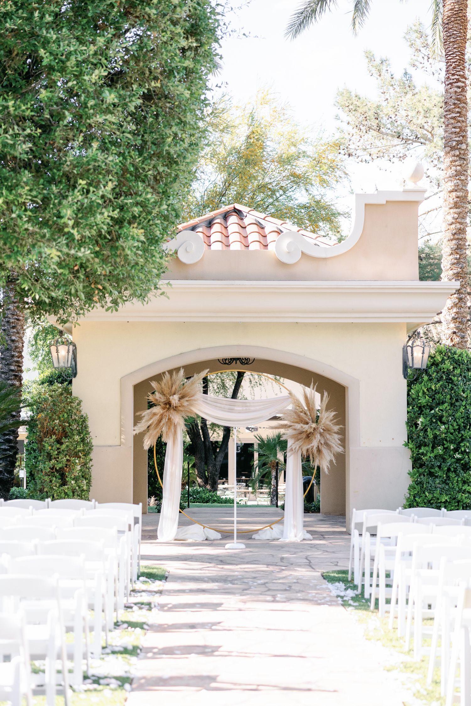 Elegant outdoor wedding ceremony setup with white chairs, draped arch, and pampas grass decor framed by lush greenery.