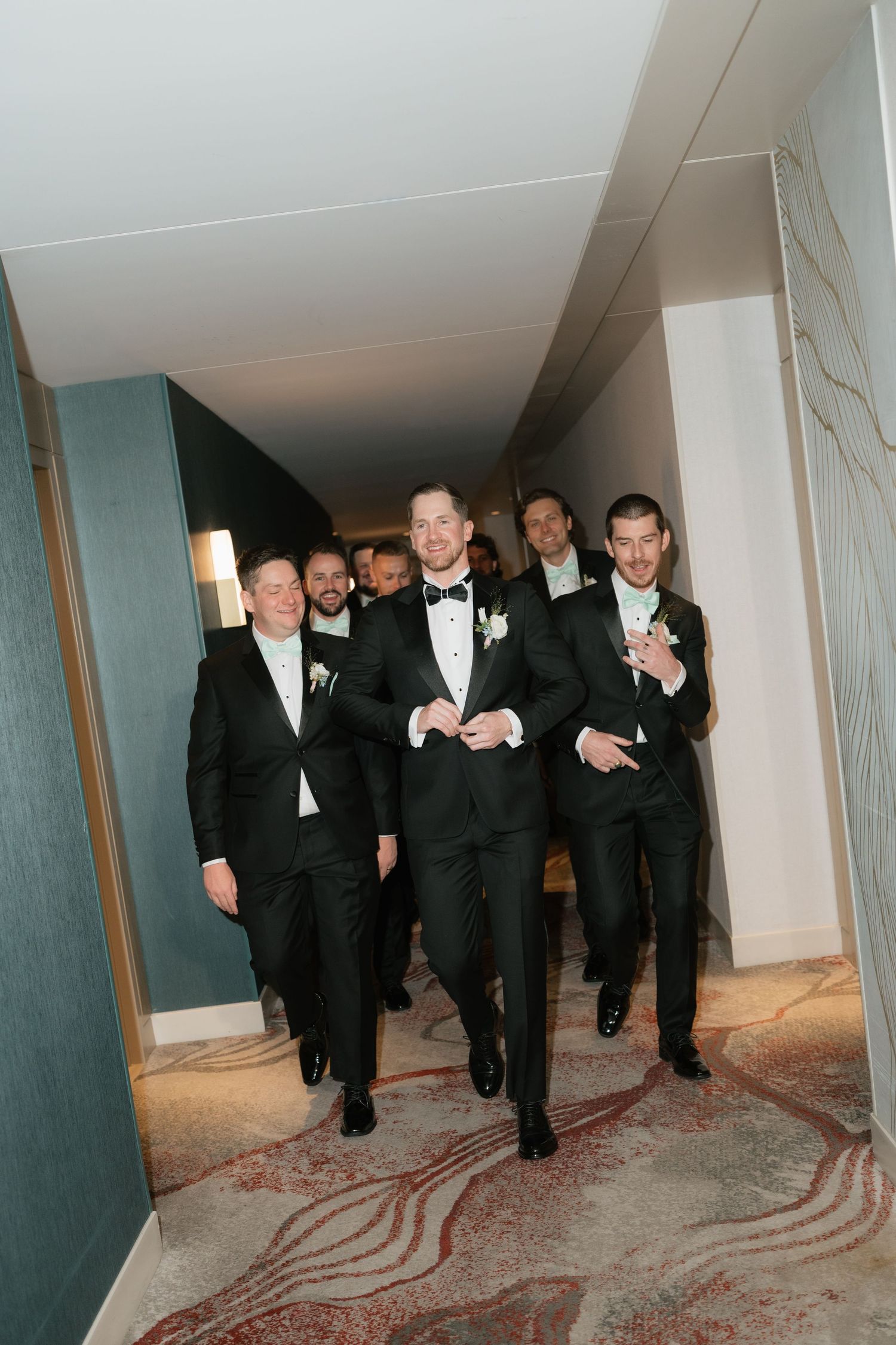 Groomsmen in black tuxedos walking confidently down a hotel corridor before a wedding ceremony.