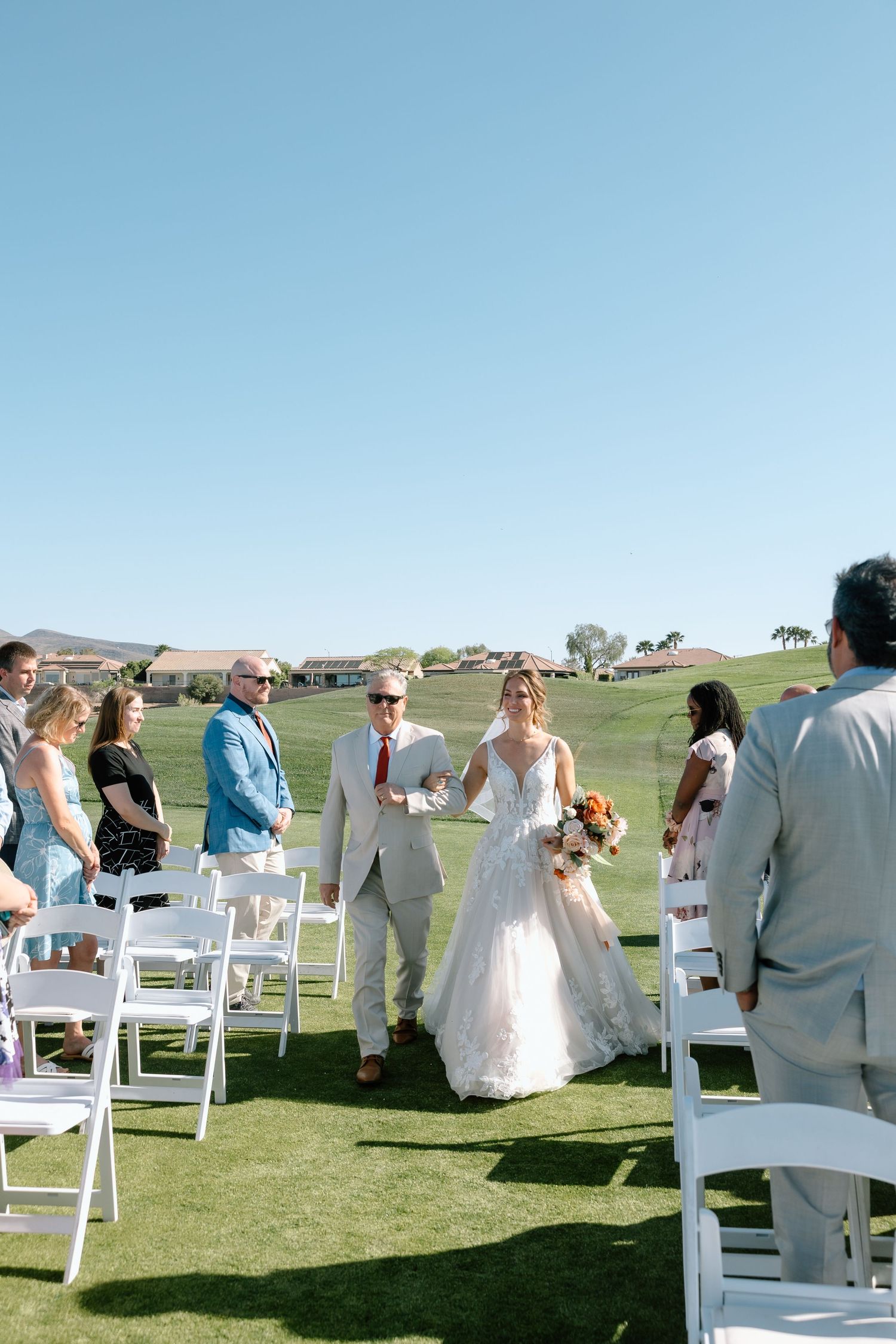 Bride walks down the aisle with escort at outdoor wedding ceremony on lush green golf course under clear blue sky.