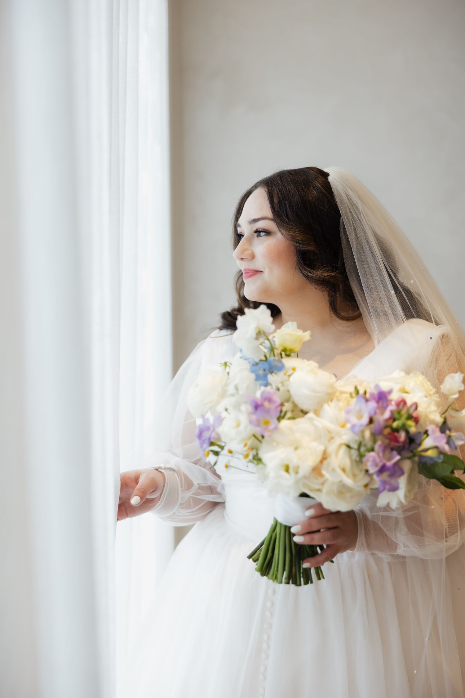 A beautiful bride in a white gown and veil holds a colorful bouquet of flowers while gazing out a bright window.
