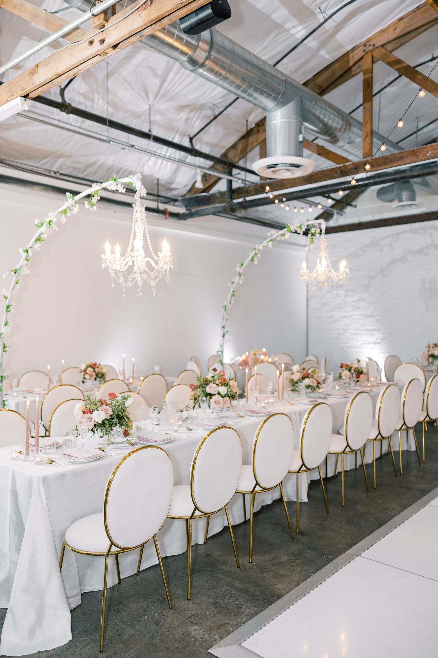 Elegant white and gold wedding reception with oval-backed chairs and crystal chandeliers in an industrial venue.