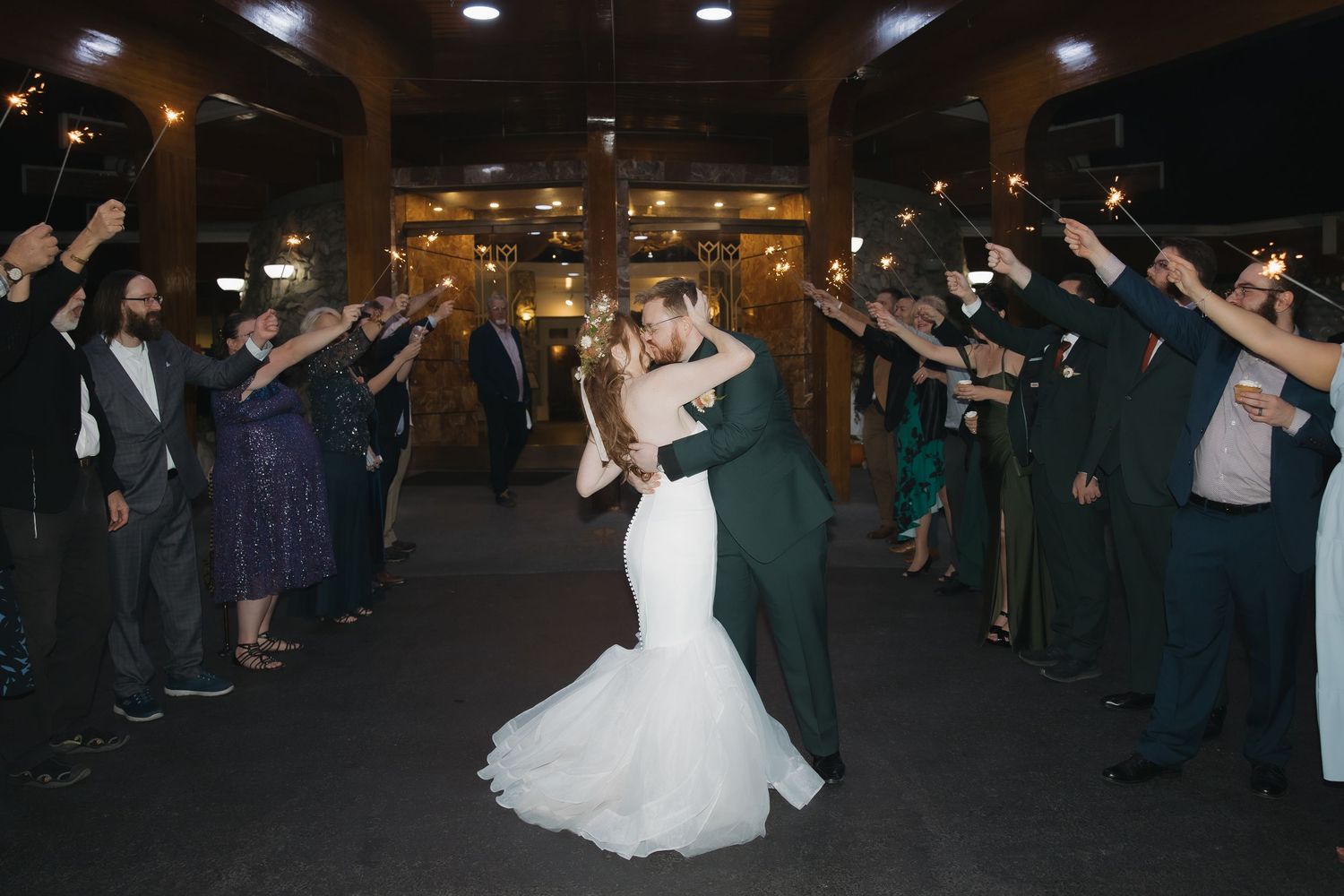 Bride and groom share a kiss during a sparkler send-off exit surrounded by wedding guests at night.