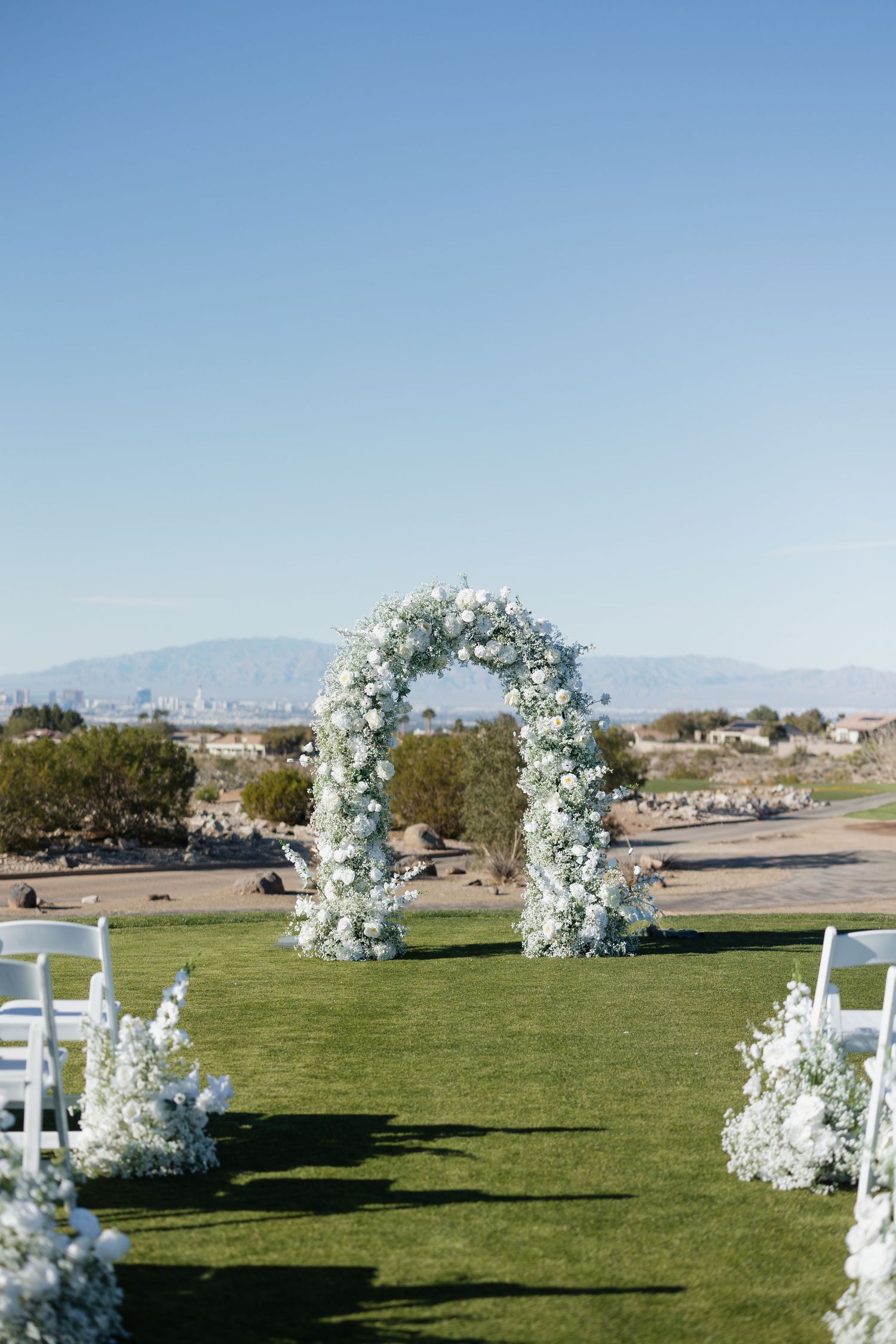 Elegant outdoor wedding ceremony arch adorned with white flowers on lush green grass, with desert mountains in the background.