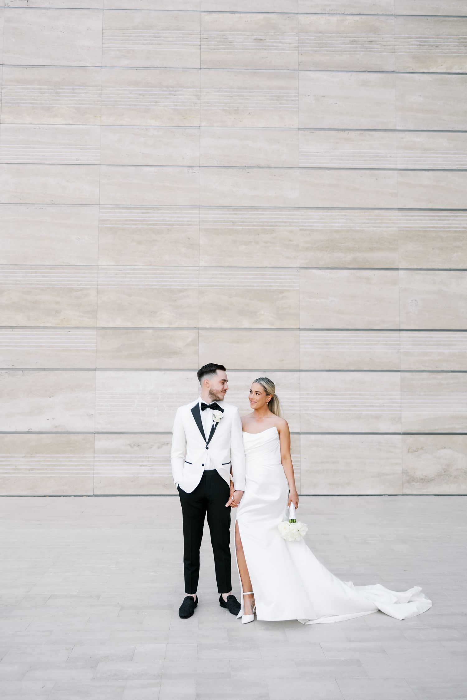 Bride and groom holding hands in front of a stone wall, wearing white wedding attire.