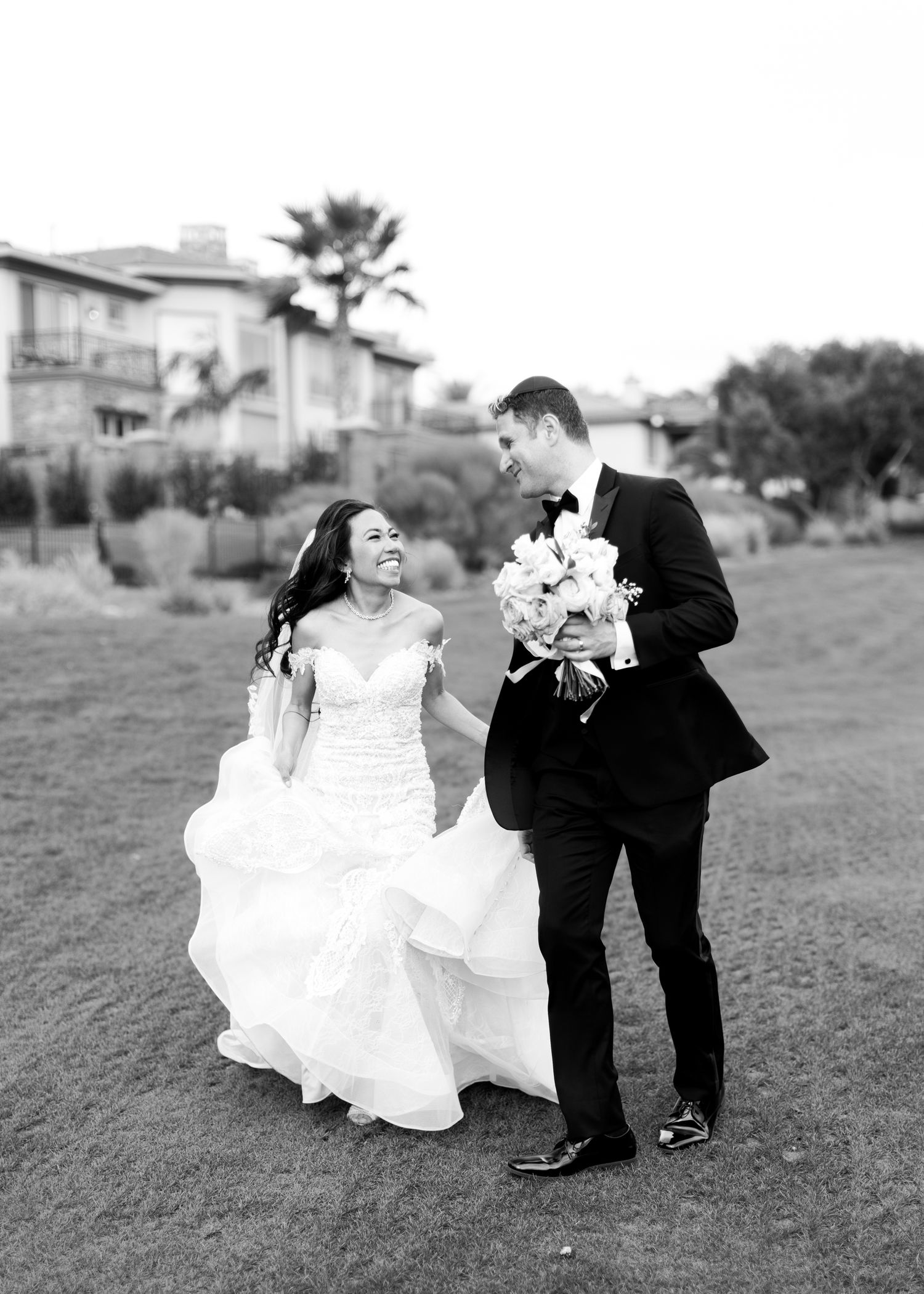 Black and white photo of a smiling bride in a ruffled gown and groom in a tuxedo walking together on a lawn.