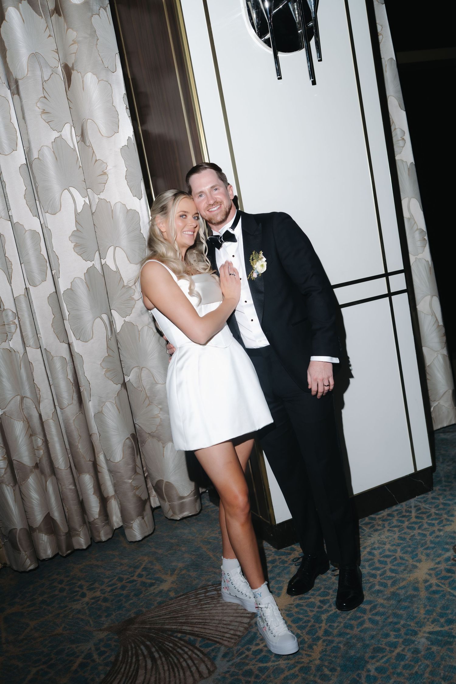 Bride in short white reception dress poses with groom in black tuxedo against floral wallpaper backdrop.
