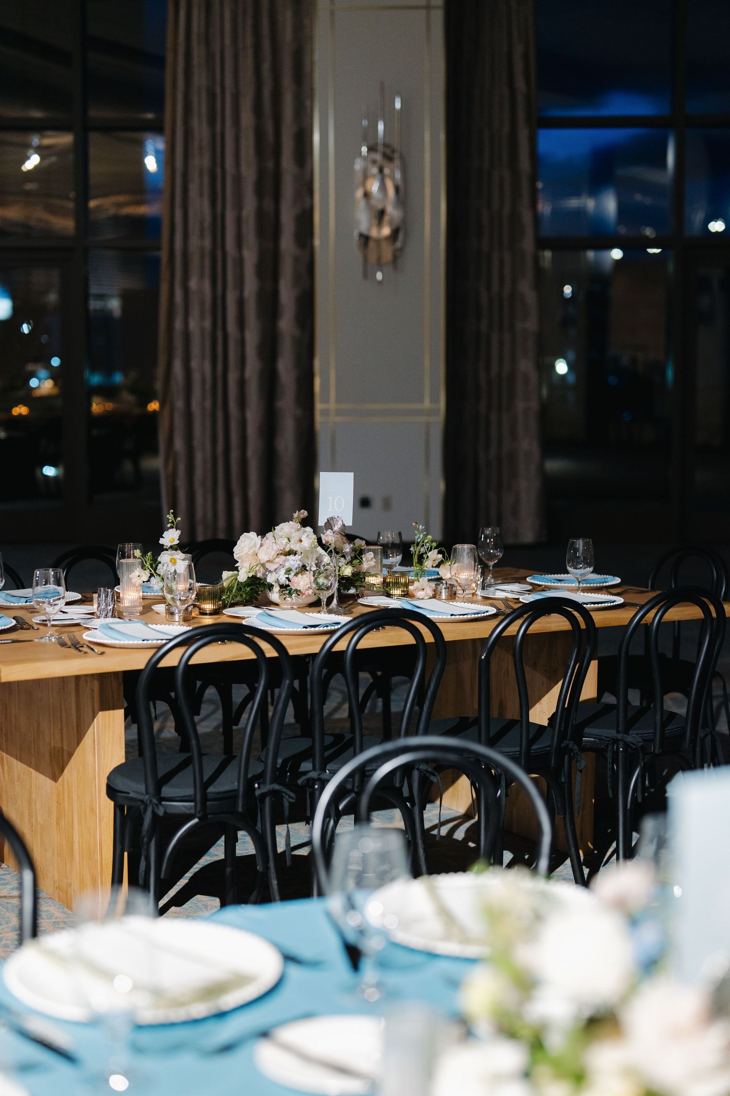 Sophisticated wedding reception setup featuring wooden tables, black chairs, white floral arrangements, and candlelight.