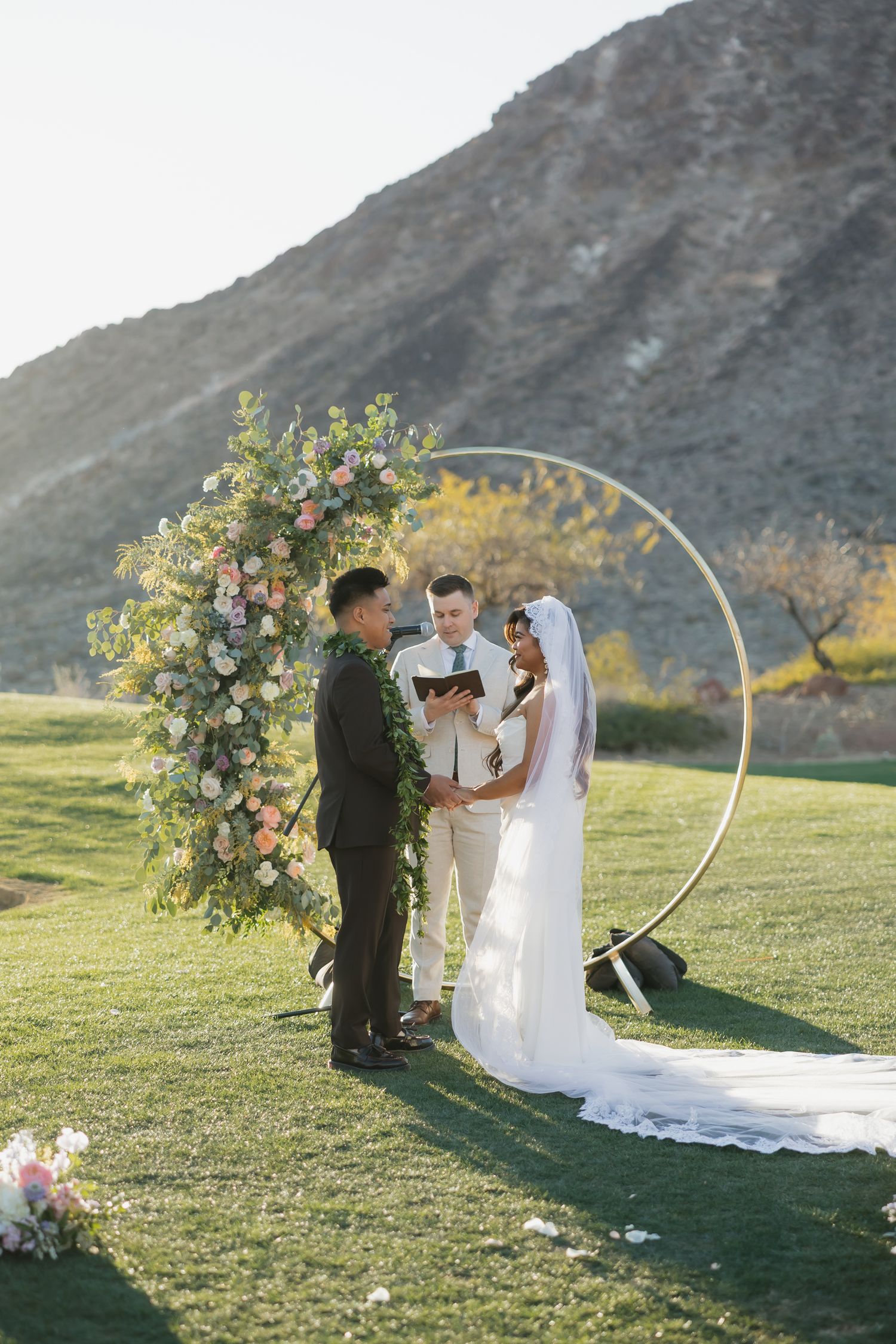 Outdoor wedding ceremony with couple exchanging vows under a circular floral arch on green grass with mountain backdrop.