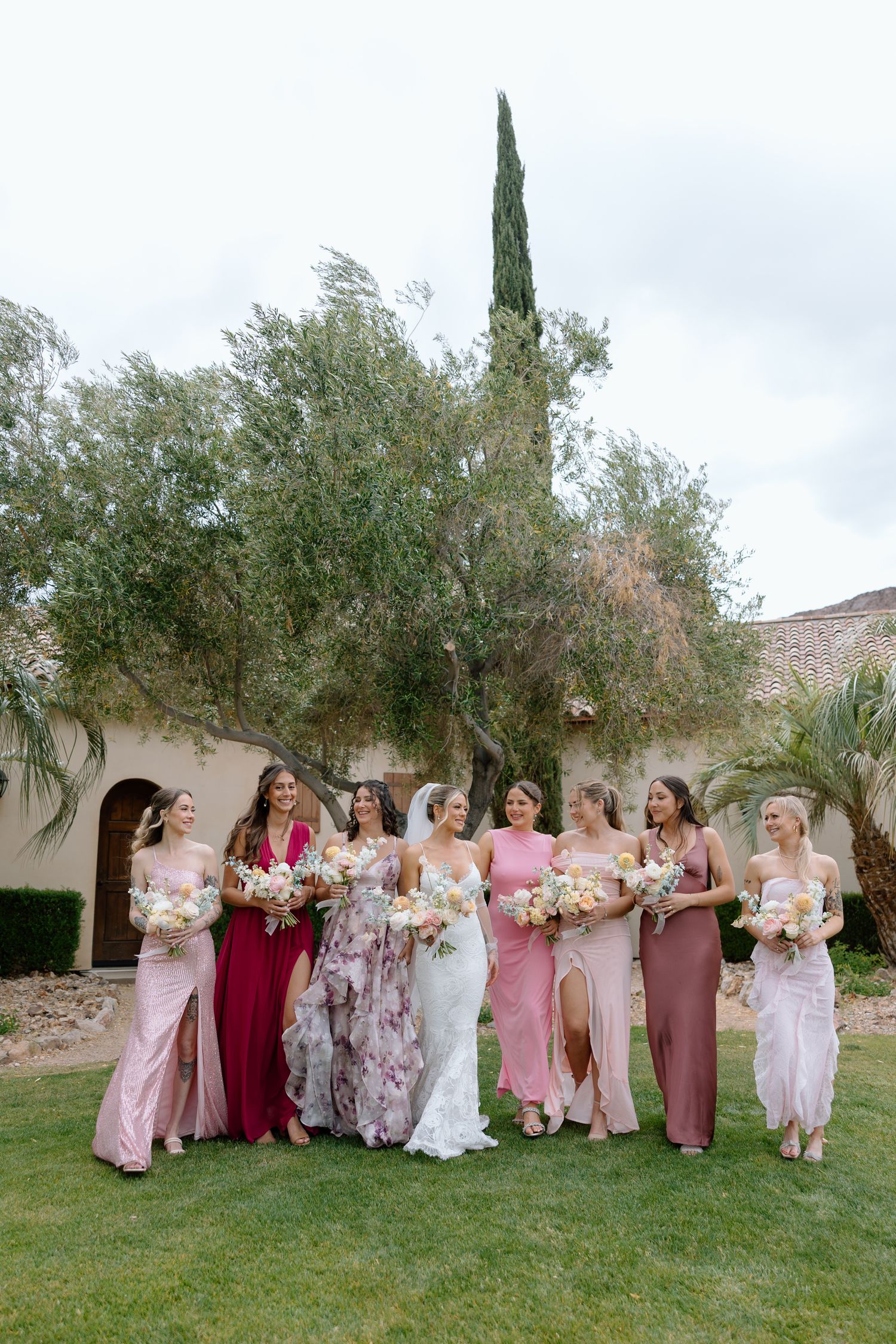 Bride and bridesmaids in pink and burgundy gowns holding bouquets outdoors at a garden wedding venue.