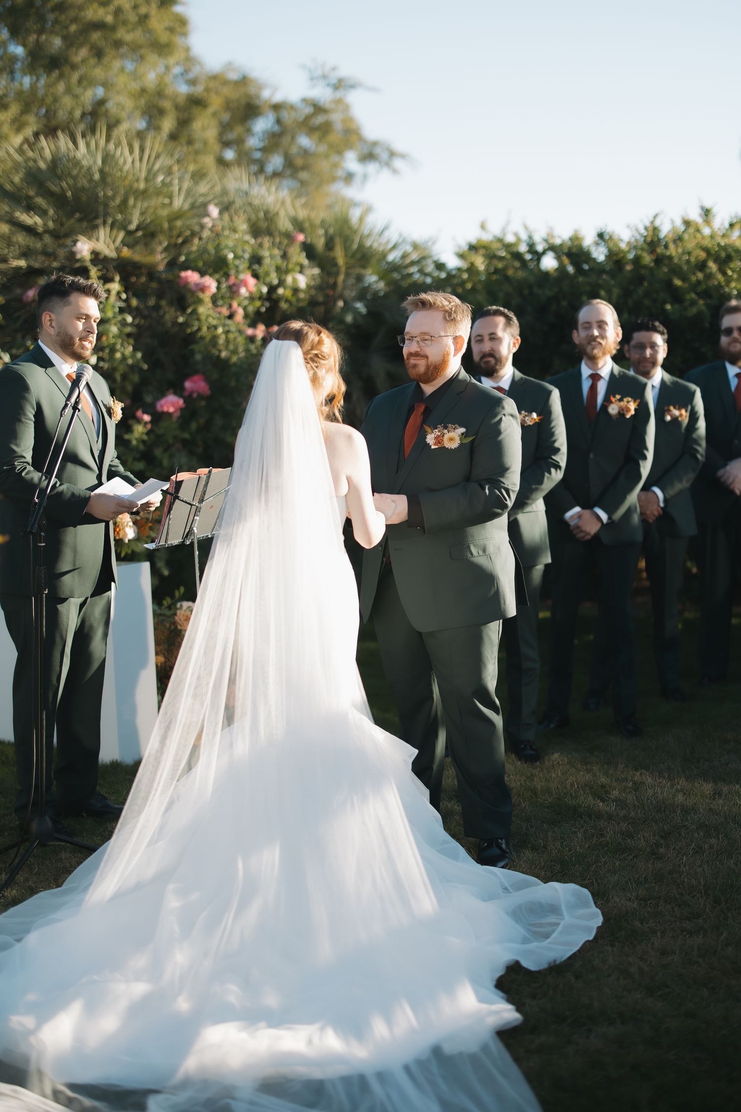 Groom in dark green suit smiles at bride in white gown and veil during outdoor wedding ceremony with groomsmen.