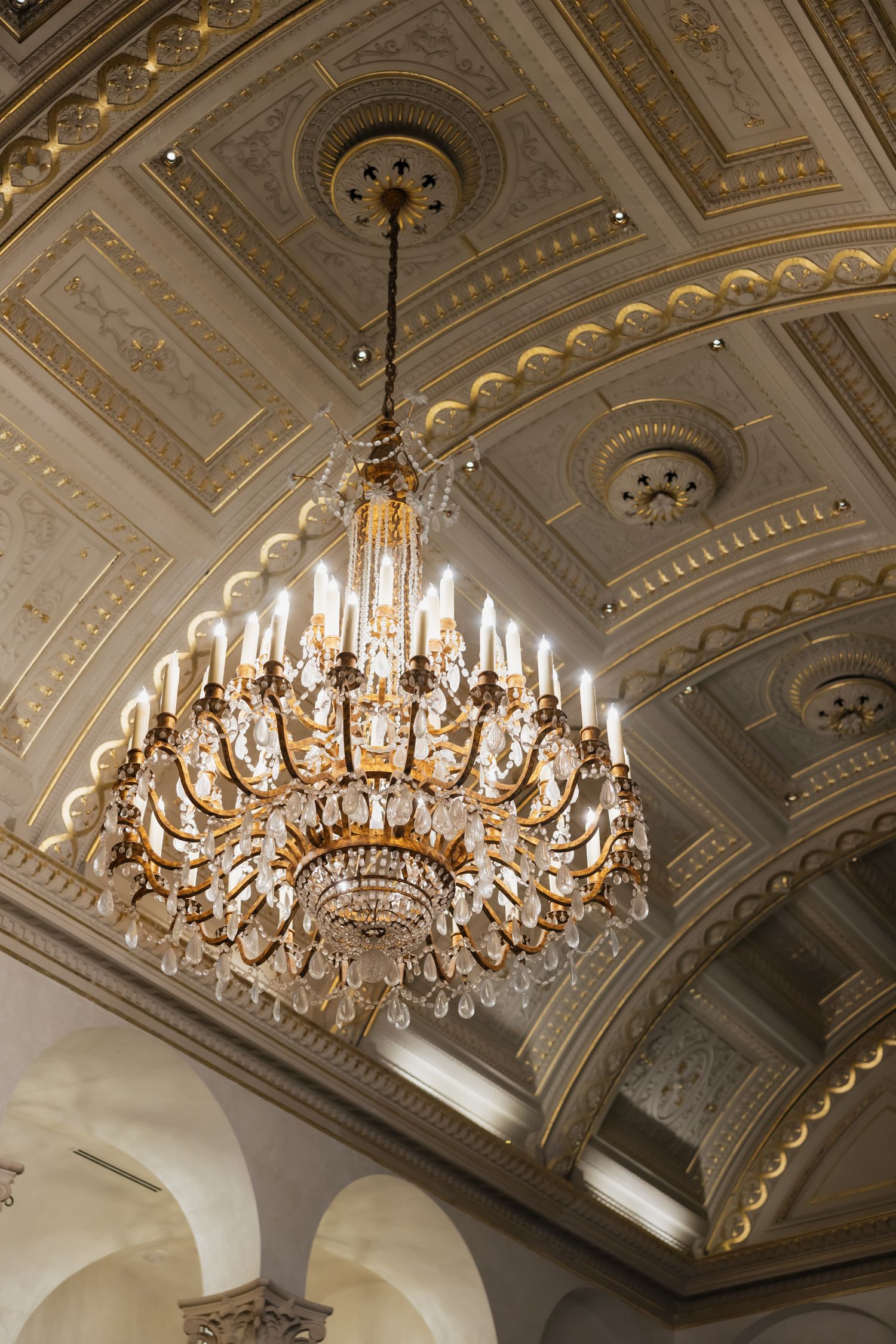 Ornate crystal and gold chandelier hanging from decorative coffered ceiling in elegant ballroom.