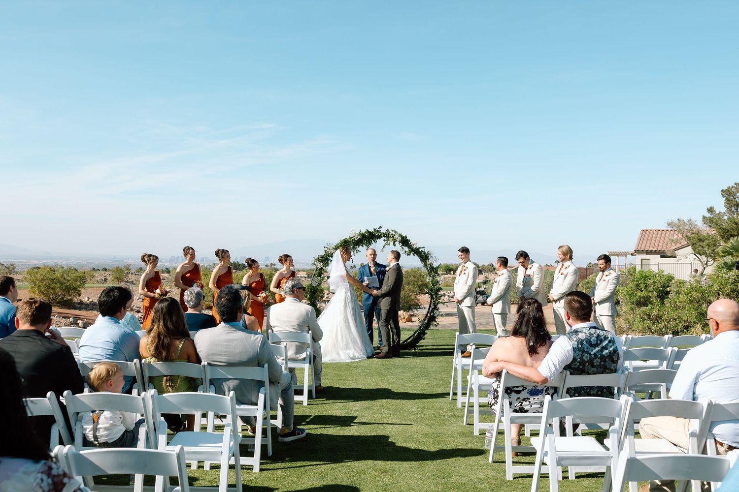 Outdoor wedding ceremony on a green lawn with white chairs, floral arch, wedding party, and guests under a clear blue sky.
