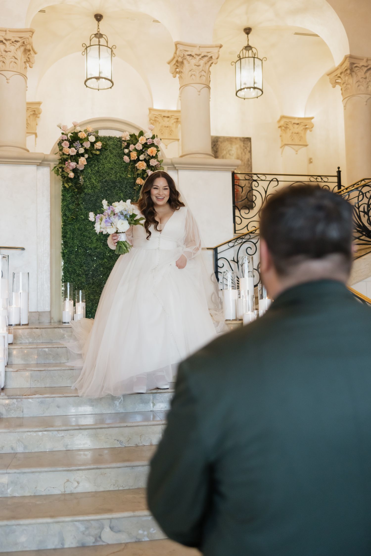 A smiling bride in a ballgown descends elegant marble stairs toward her groom at a grand wedding venue.