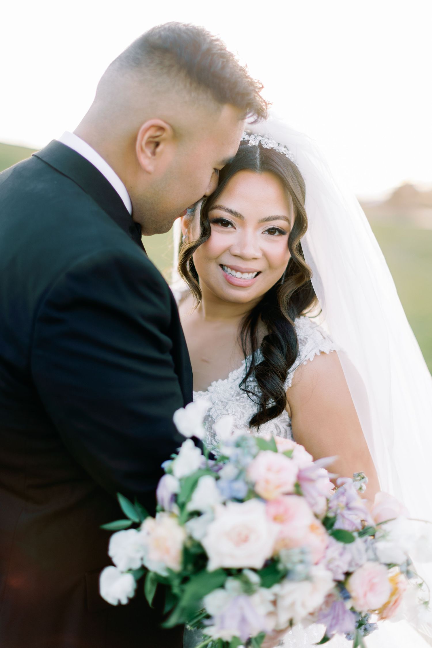 Bride smiling with groom nuzzling her cheek, holding a pastel floral bouquet in soft golden light.