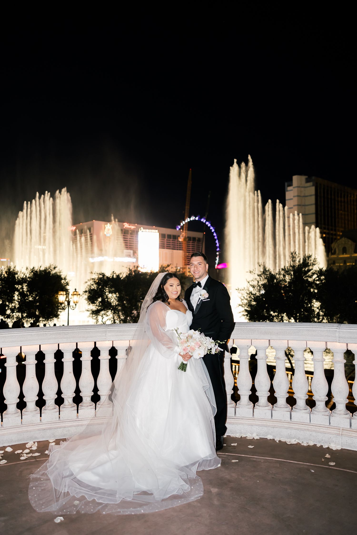 Bride and groom pose by Bellagio fountains at night in Las Vegas, bride holding white bouquet in elegant gown.