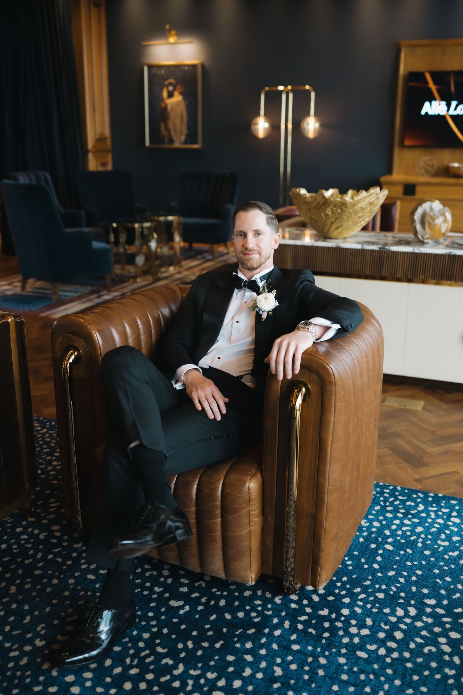 A groom in a black tuxedo with boutonniere sits confidently in a brown leather chair in an elegant hotel lounge.