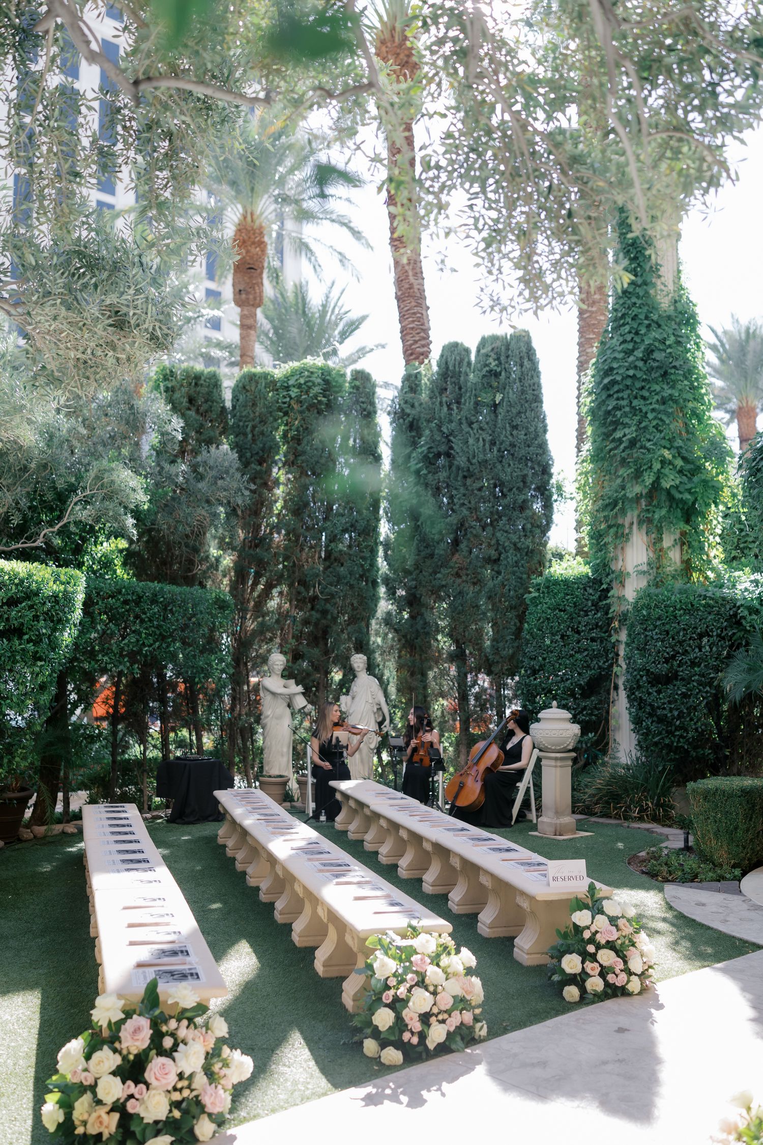 A row of white benches and floral arrangements line a garden path with palm trees and greenery creating an elegant aisle.