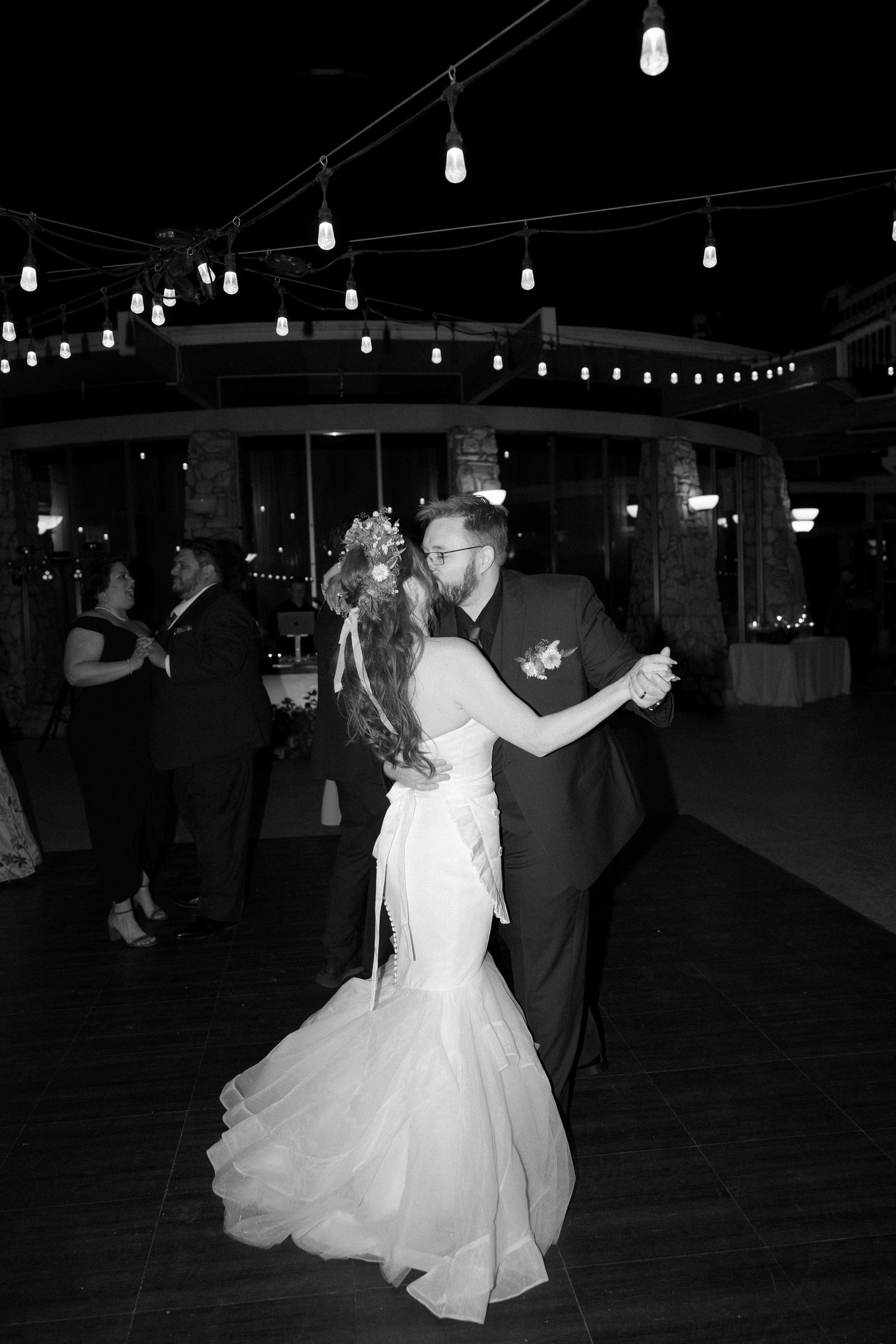 Bride and groom share first dance in black and white photo under string lights at reception.